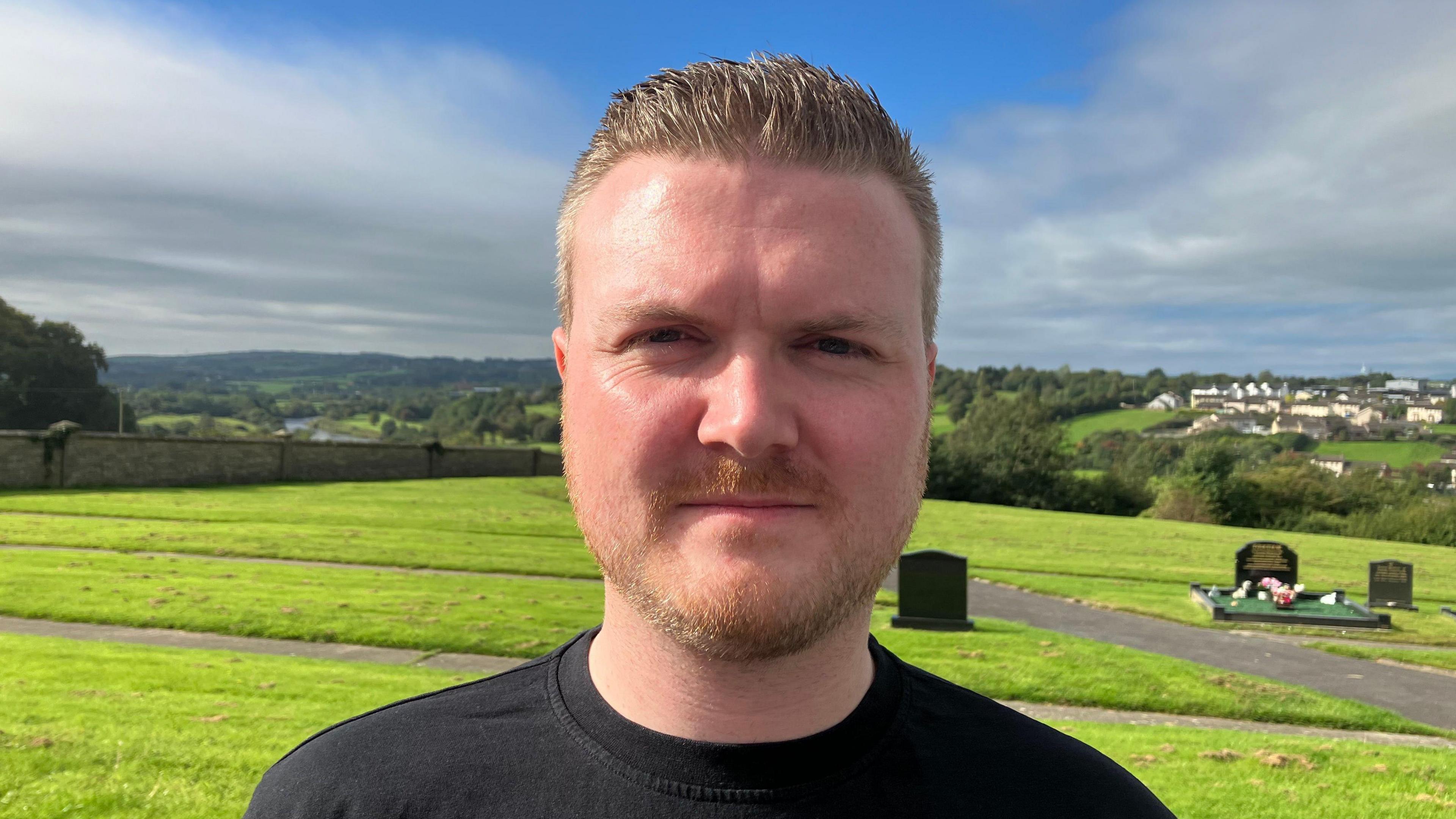 Conor Logue. He is standing in a graveyard and wearing a black t-shirt. He has light hair and a beard.