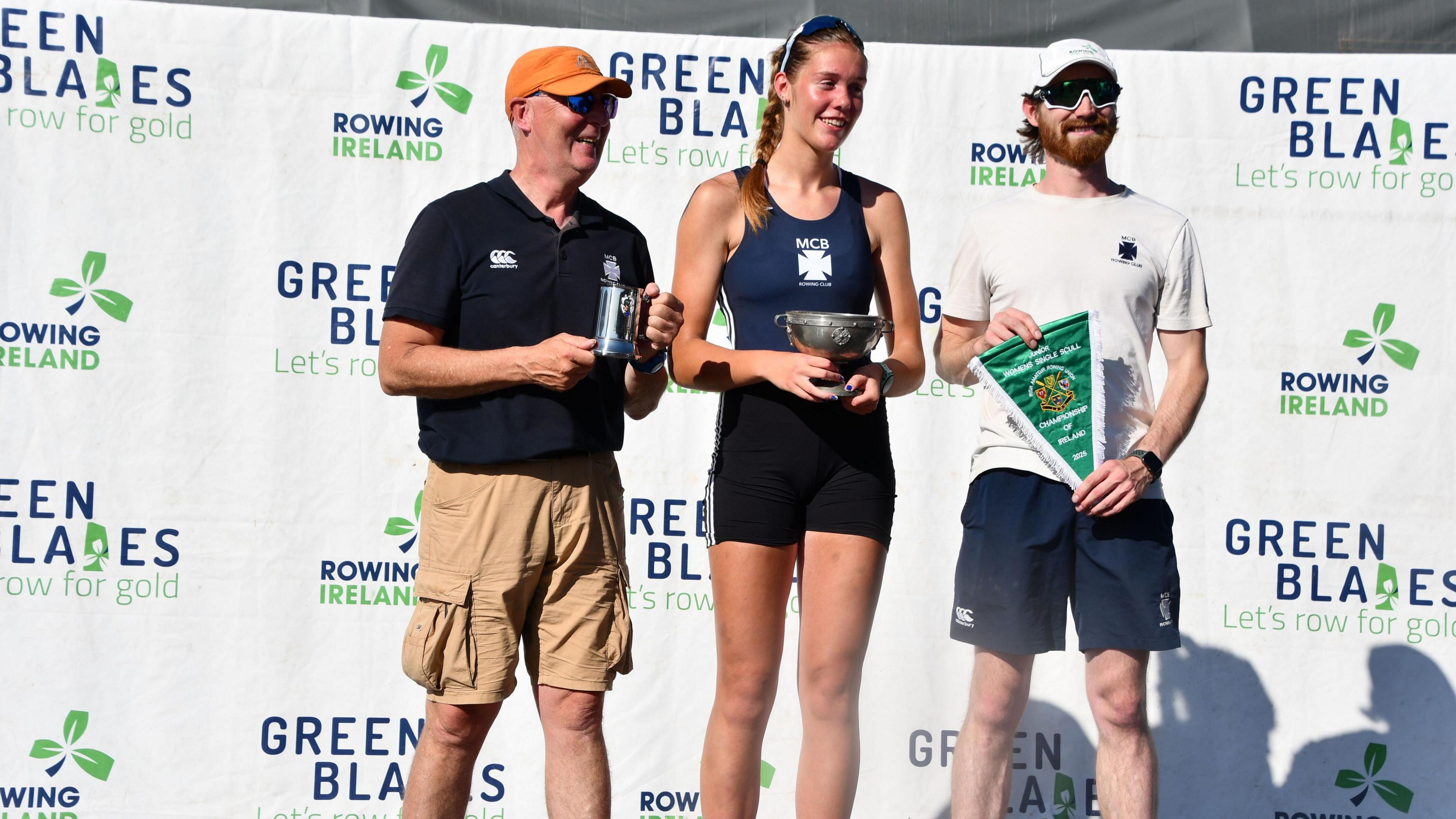 Sophia Young standing in front of a white tarped podium with her coaches. She is wearing a navy swimsuit with a white Methodist College Boat Club badge in the middle. She is holding her trophy for the event. She is flanked either side by her coaches Enda Marron and Miles Taylor.