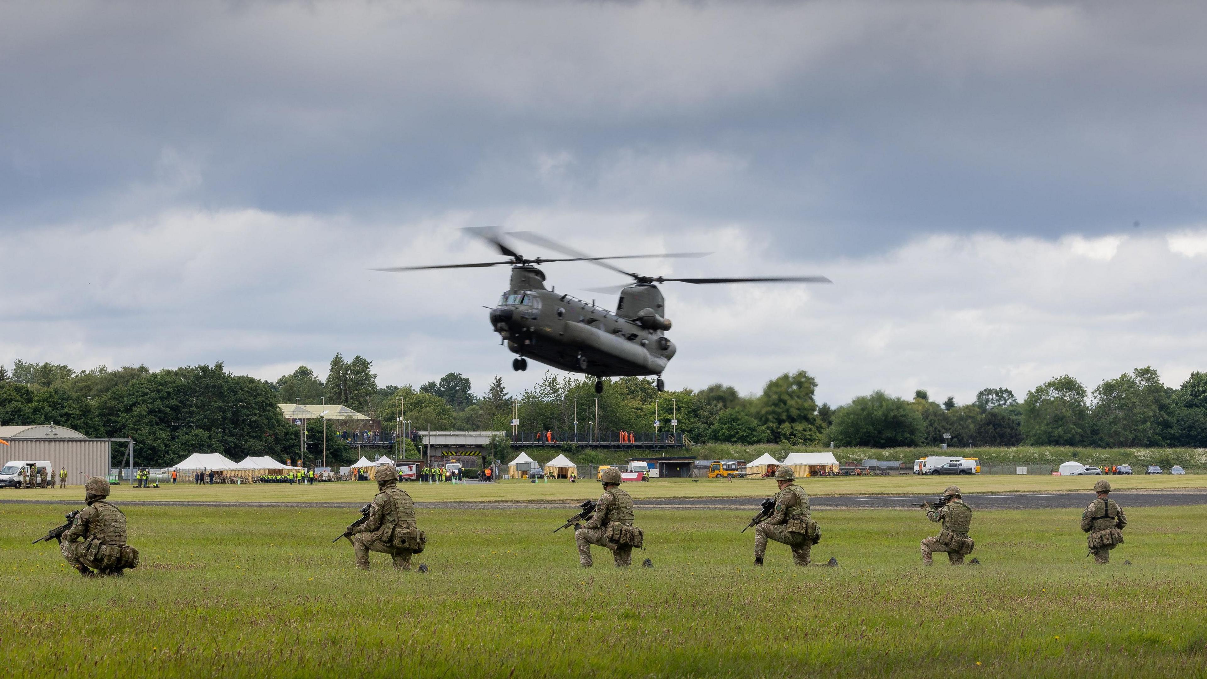 A military helicopter with twin rotors hovering low above a field. Kneeling in front of it are six soldiers in camouflage and holding guns.