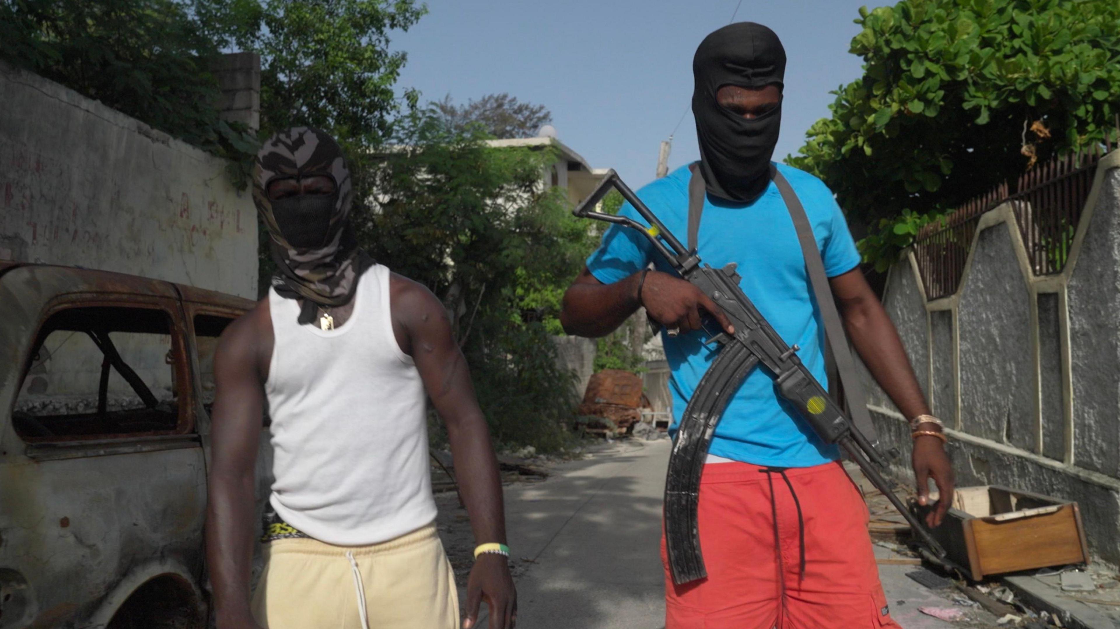 Two young men with their heads and faces covered by black hoods and masks, except for their eyes, facing the camera. The one on the right is wearing a blue T-shirt and red shorts, and carrying a large gun. The one on the left is in a white vest and pale yellow shorts.