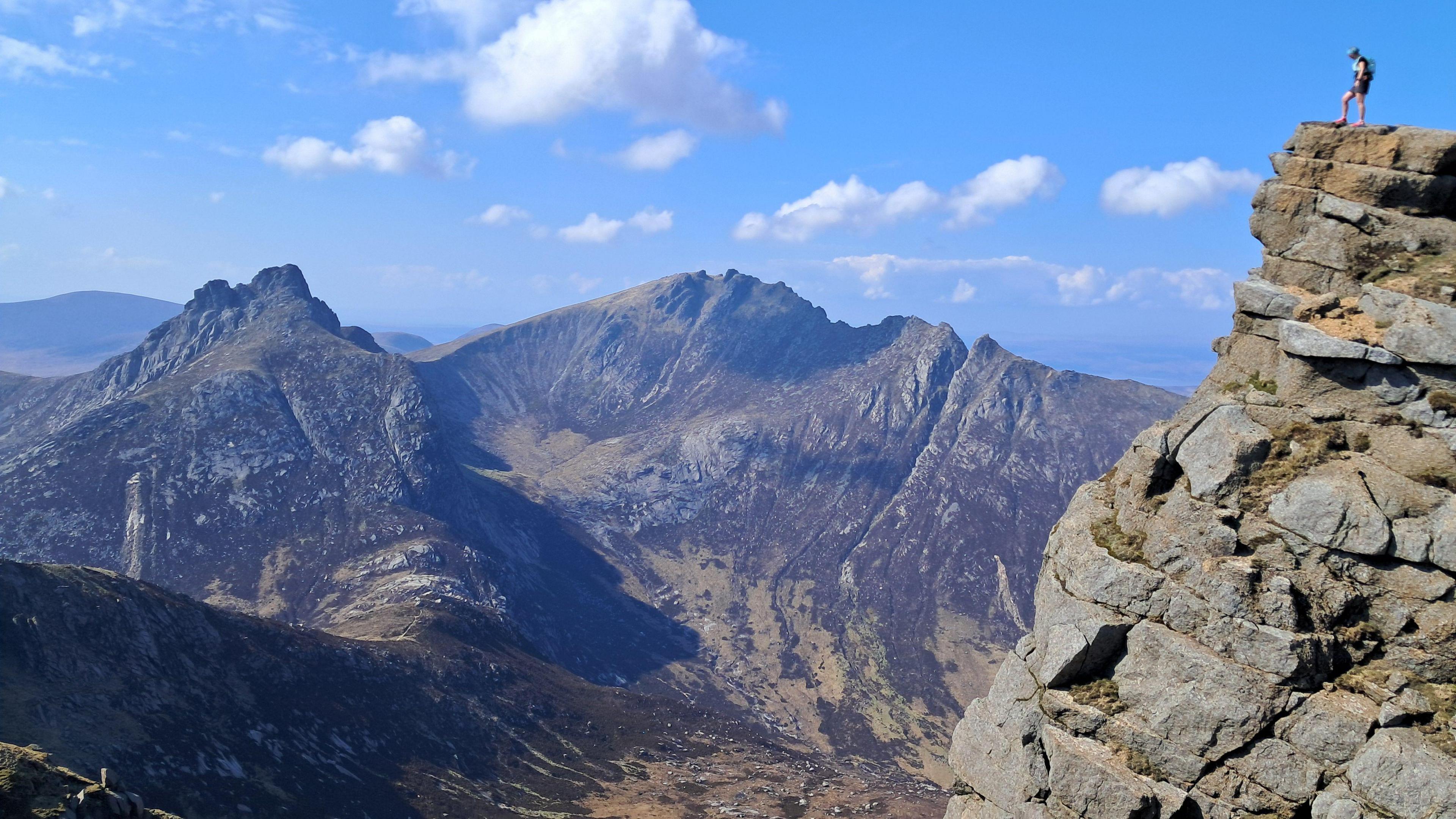 A woman stands on top of a stack of rock looking out to rocky mountains. The sky is blue.