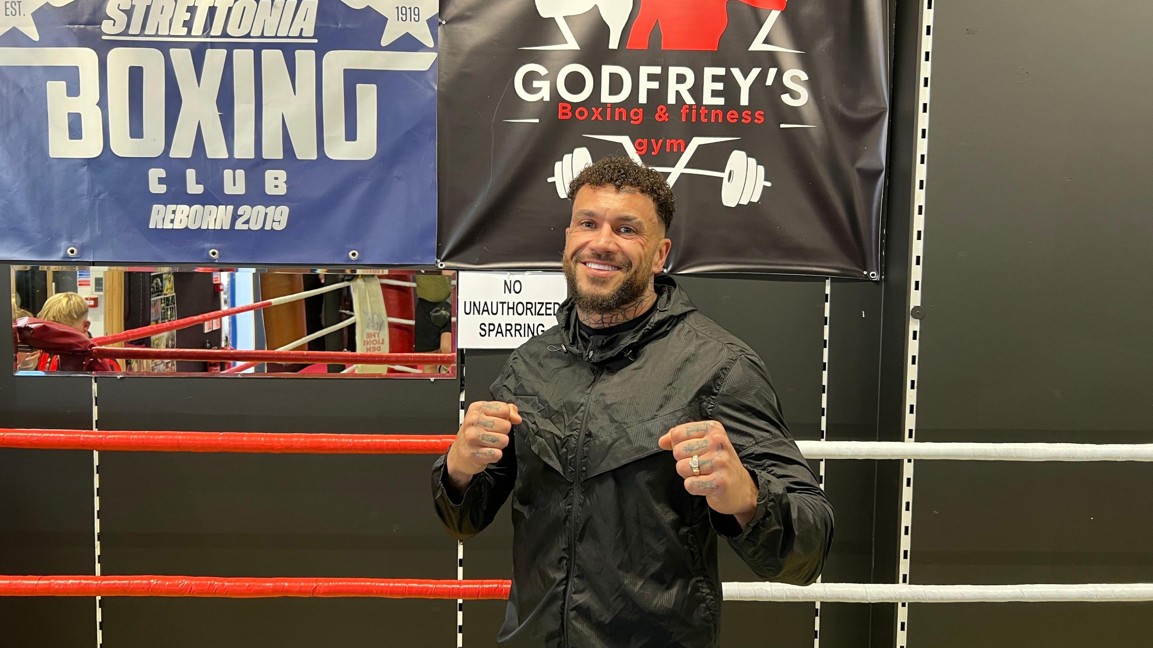 Darren has curly brown hair and brown facial hair. He has tattoos on his neck and hands. He is posing with both fists out, and is wearing a black waterproof jacket.