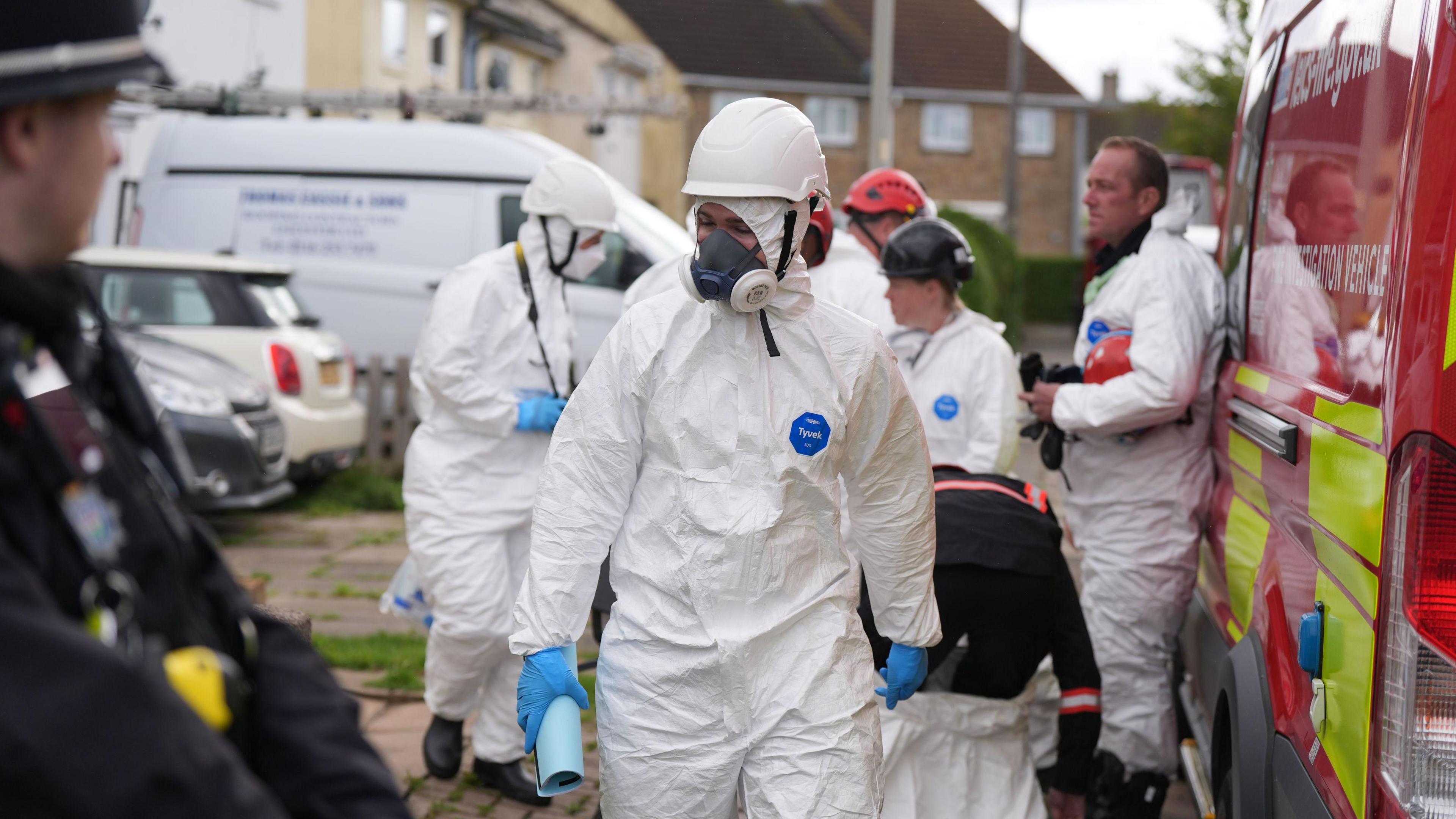 Four forensic officers in white overalls, helmets and masks next to a red fire service van 