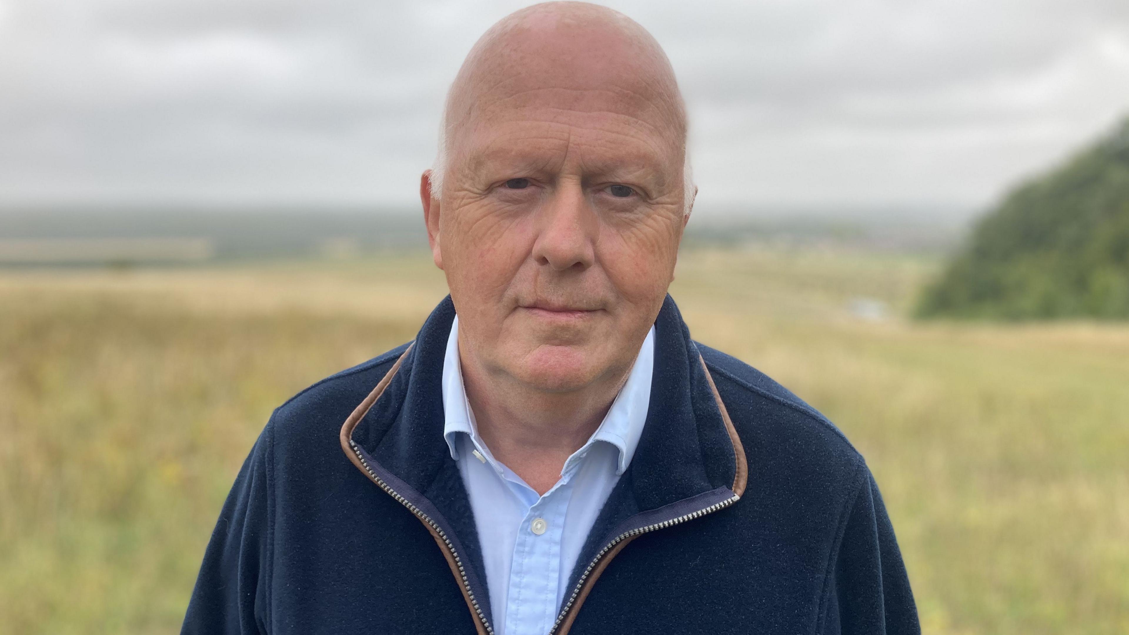 Hugh Carter standing on Salisbury Plain. he is mostly bald and wearing a blue zip-up top and light blue shirt