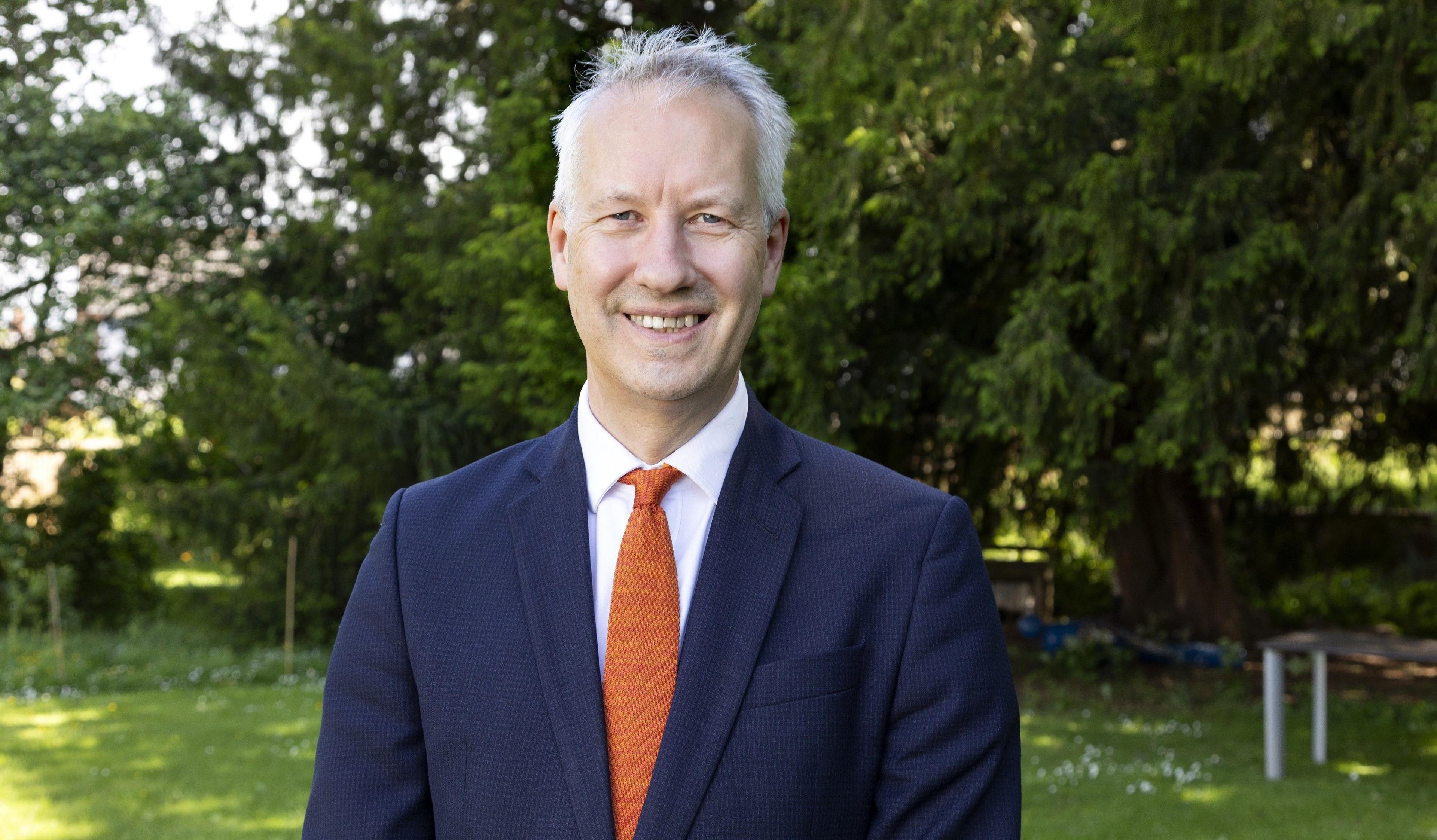 A man wearing a blue suit with an orange tie outside in the sunshine. 