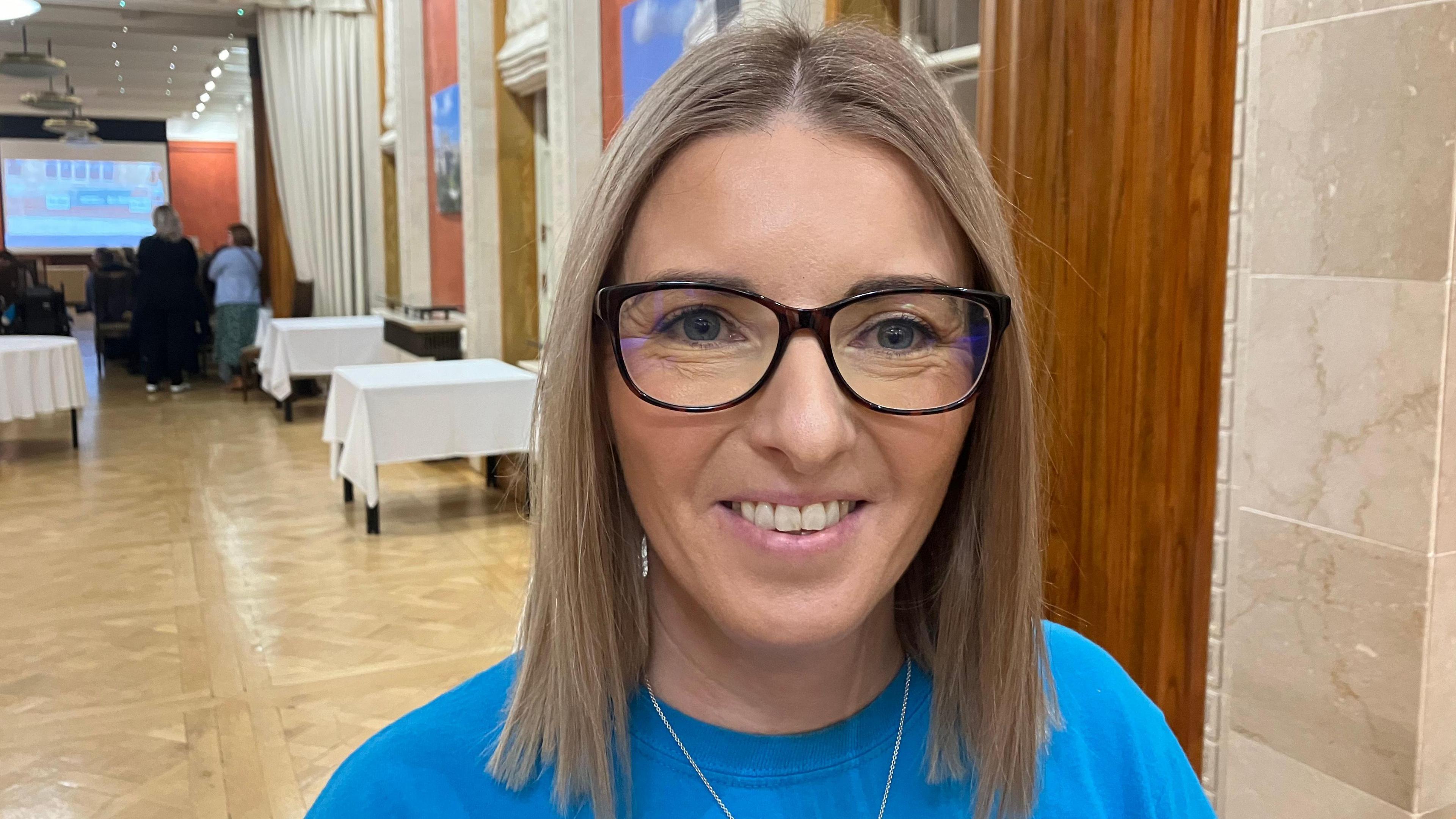 Jaclyn Harron - a women with shoulder-length blonde hair stands in a large hall. She is wearing black, square-rimmed glasses and a blue shirt. She is smiling at the camera.