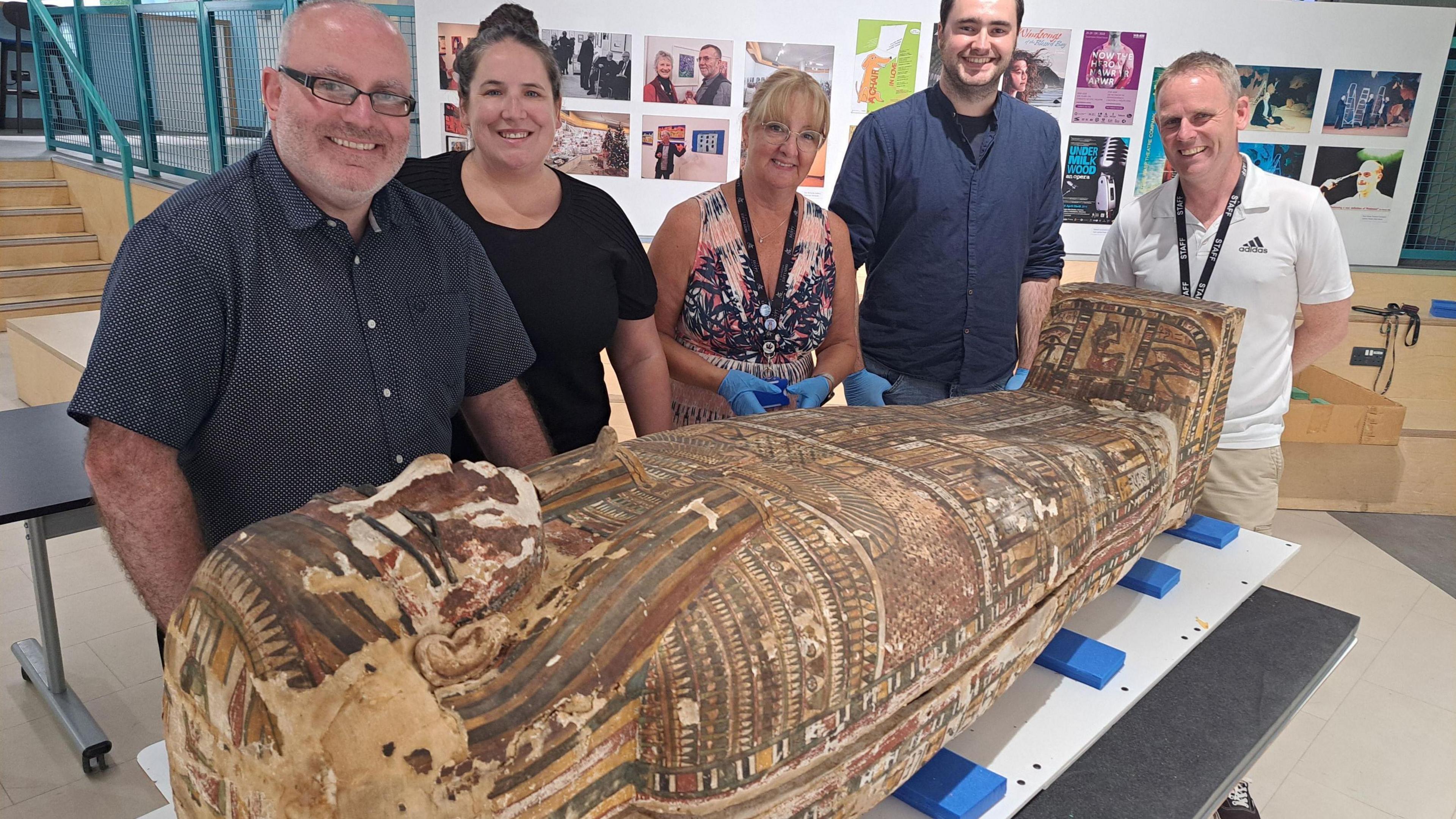 Five staff at Swansea University smiling as they pose with the ancient Egyptian coffin