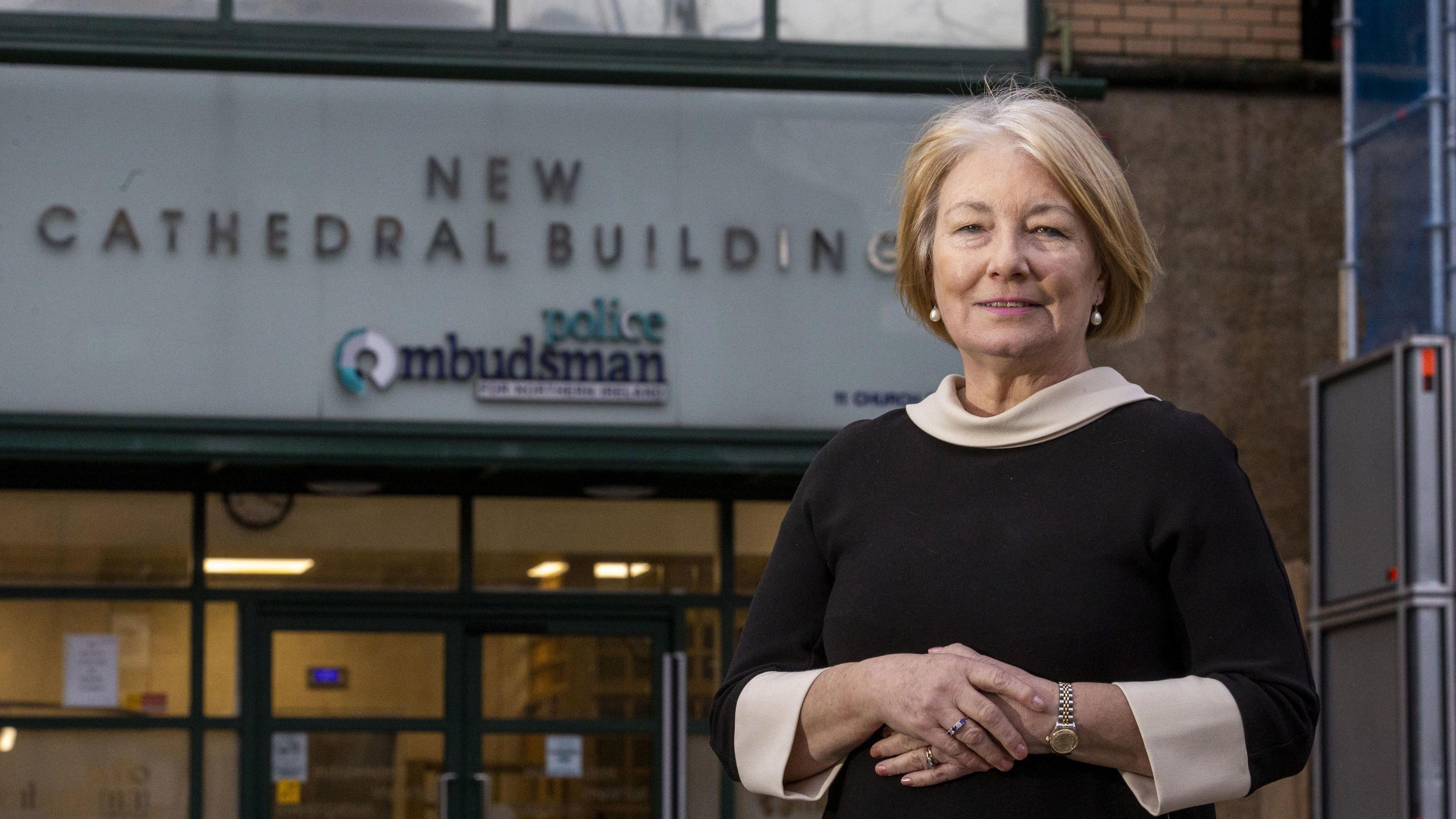 Marie Anderson. She has a blonde bob, wearing pearl earrings, a black dress with cream cuffs and neckline. She is standing in front of a with a sing on it saying 'New Cathedral Building Police Ombudsman' 