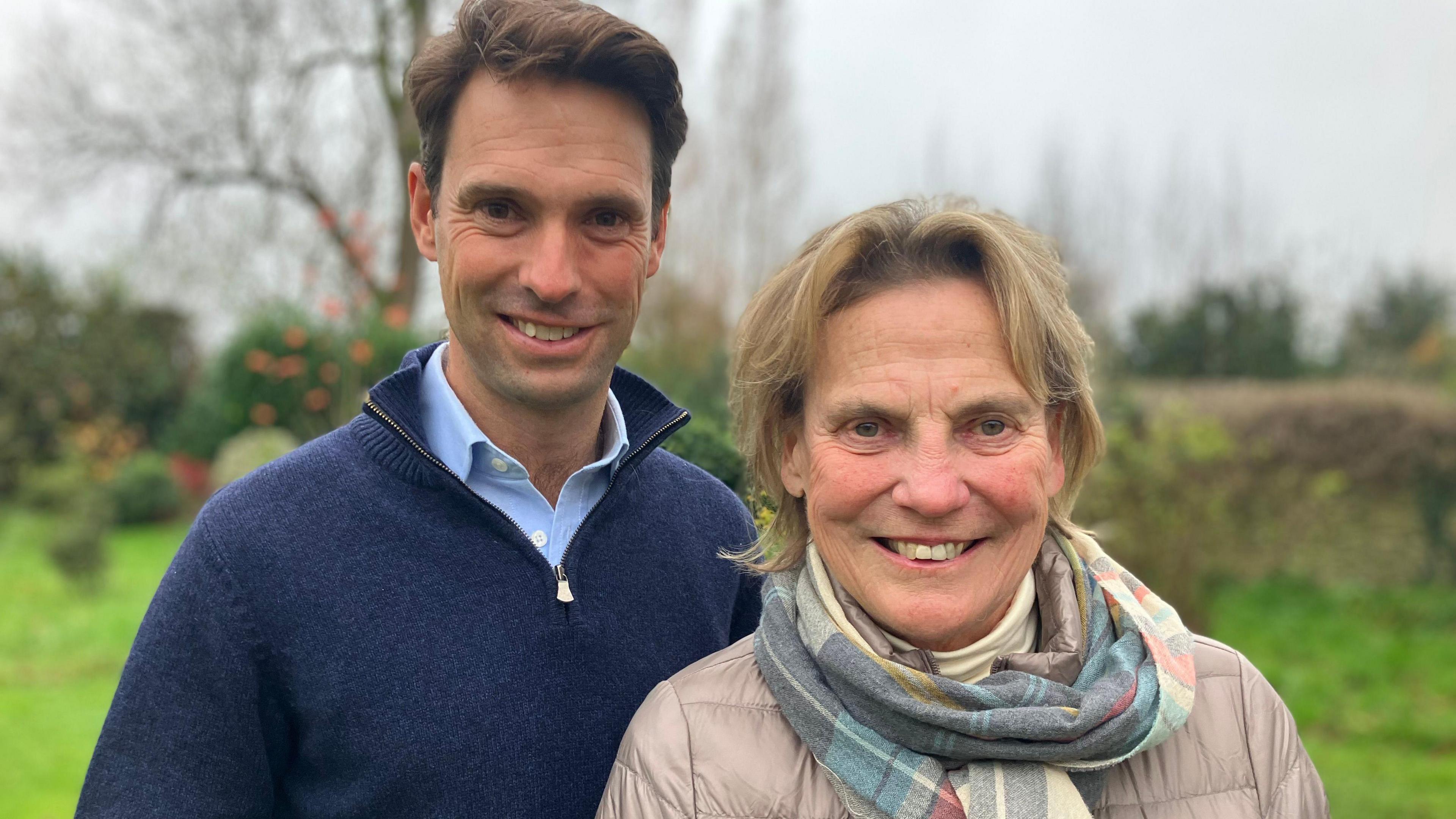 A man wearing a blue jumper stood next to a woman in a grey jacket and blue and grey scarf. They are stood in a garden and behind them are blurred hedges and a tree. 