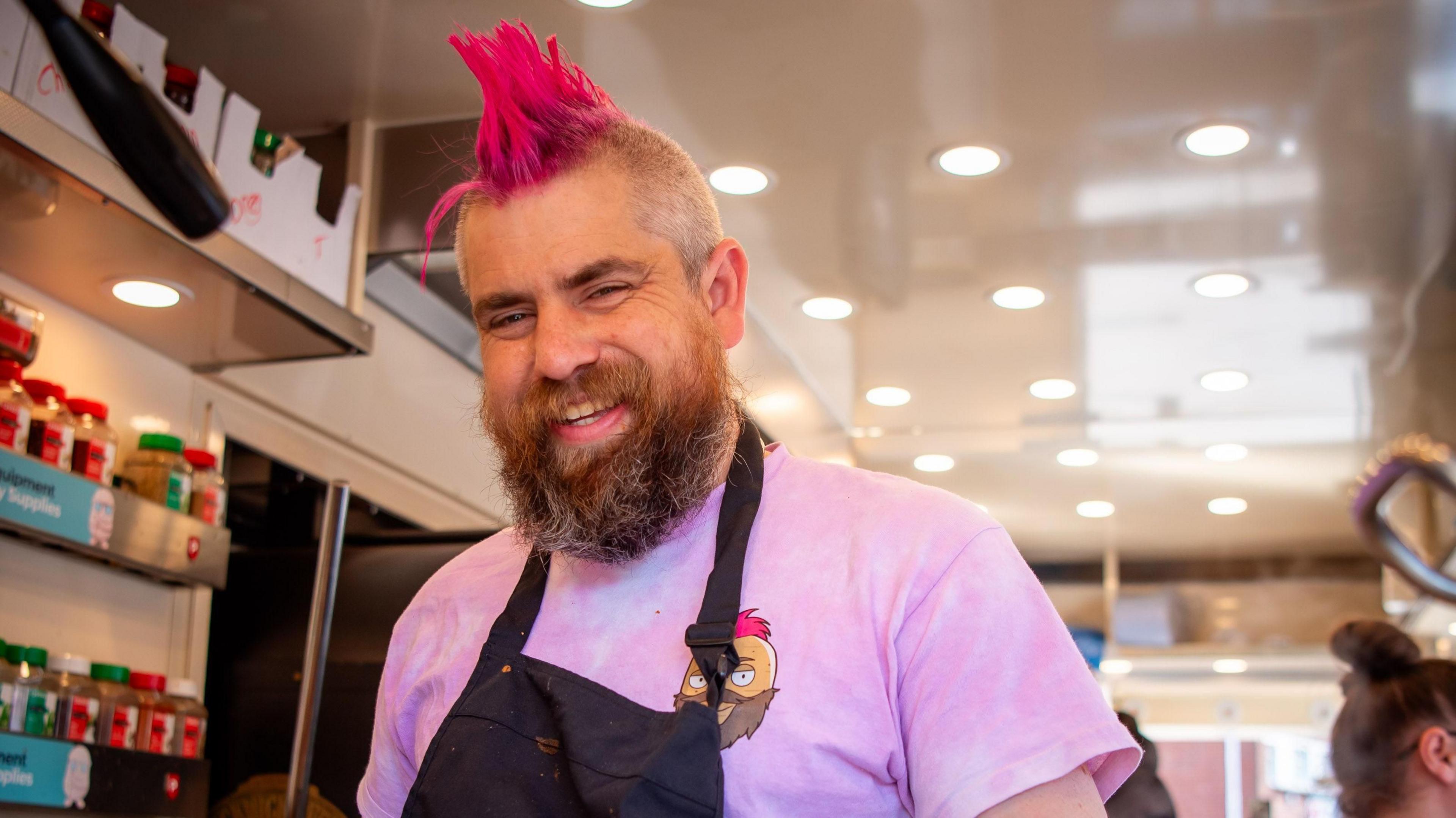 A man with a beard and pink mohawk hair is wearing a pink T-shirt and black apron. He is smiling and standing in a brightly-lit room with herb and spice jars on shelves next to him.