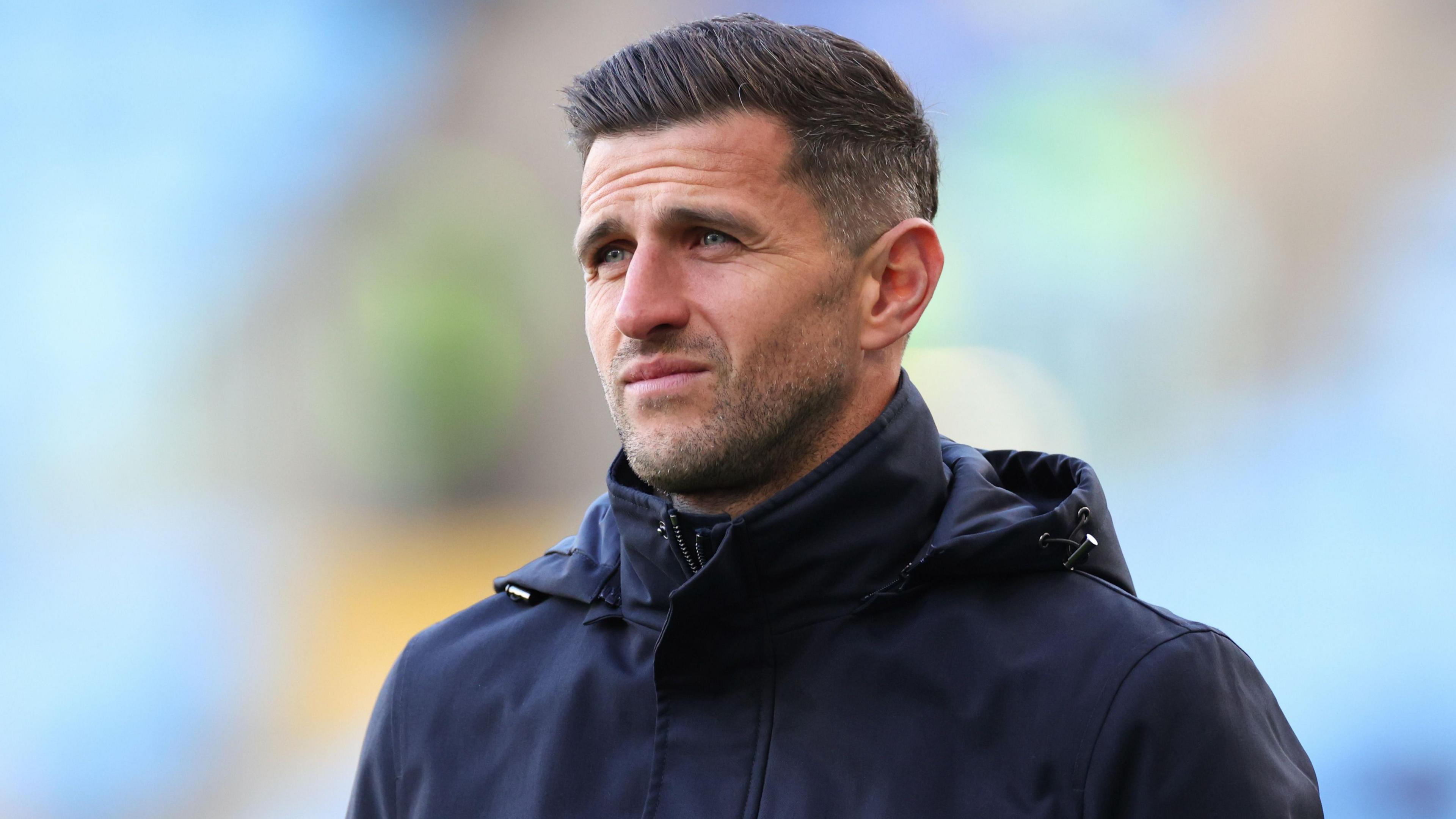 John Mousinho, wearing a black coat, standing on the sideline in the 5-1 loss at Coventry 