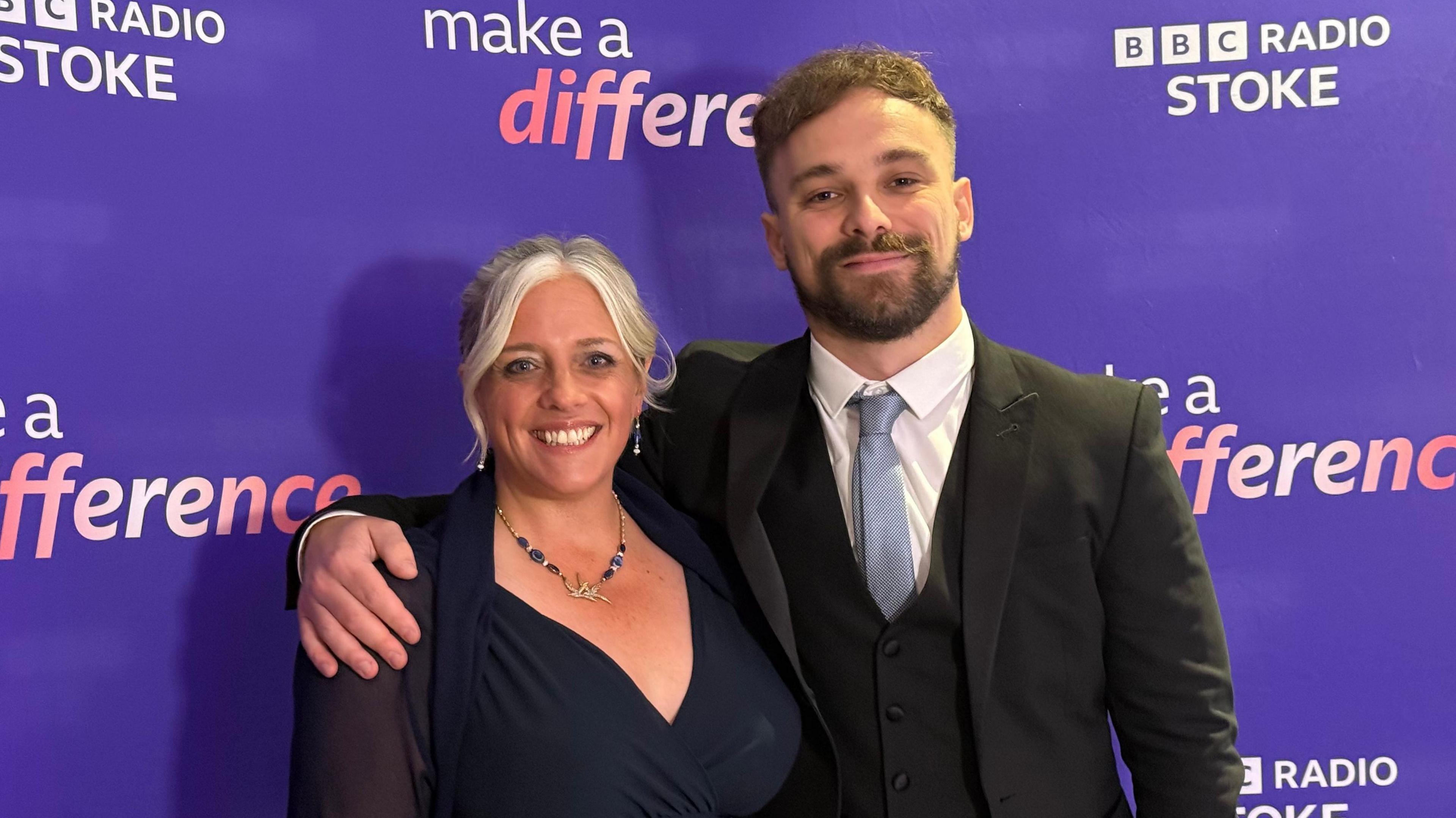 Jodie Looker and Lee Blakeman - Jodie is wearing a dark blue dress and Lee Blakeman is wearing a black suit with pale blue coloured tie and white shirt. They are stood against a purple BBC Radio Stoke Make A Difference step and repeat board.