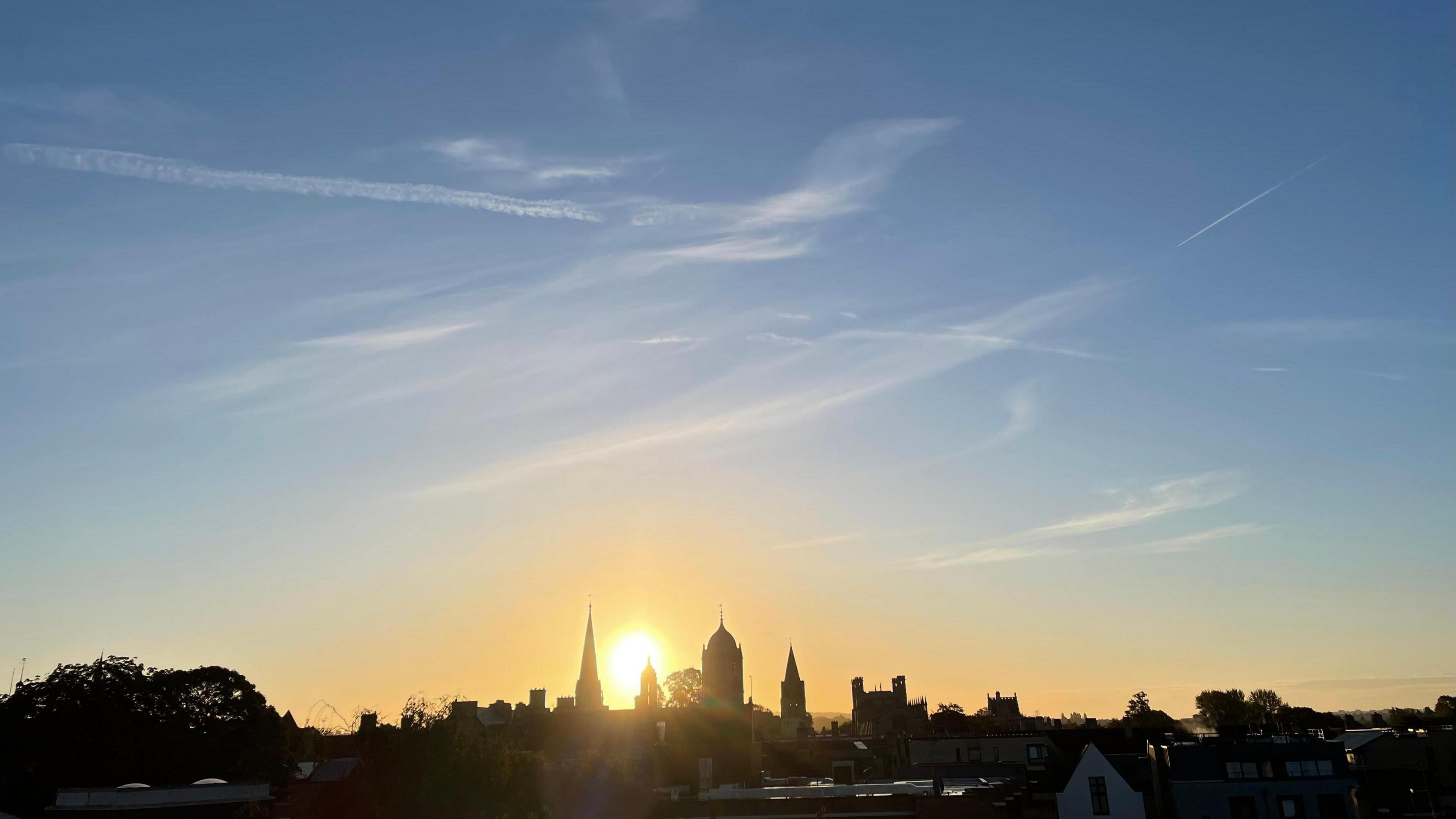 An early morning as the sun rises above the Oxford skyline. The building son the horizon are in silhouette with a bright sun just rising above them. The sky is pale blue with some small wispy clouds and contrails criss-crossing it.