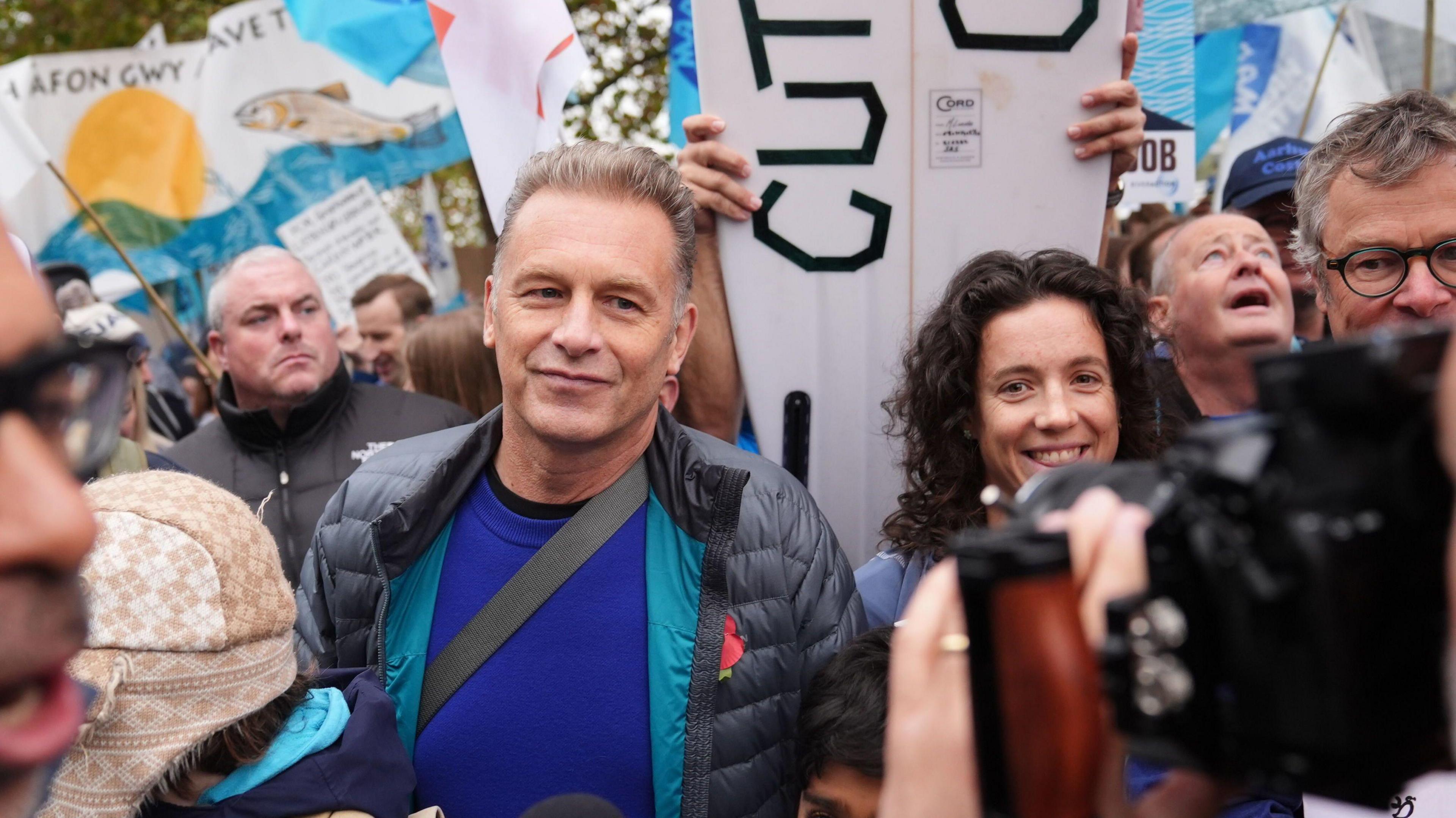 Chris Packham in a crowd. He is wearing a blue top with a black coat on top. People behind him are holding up banners.