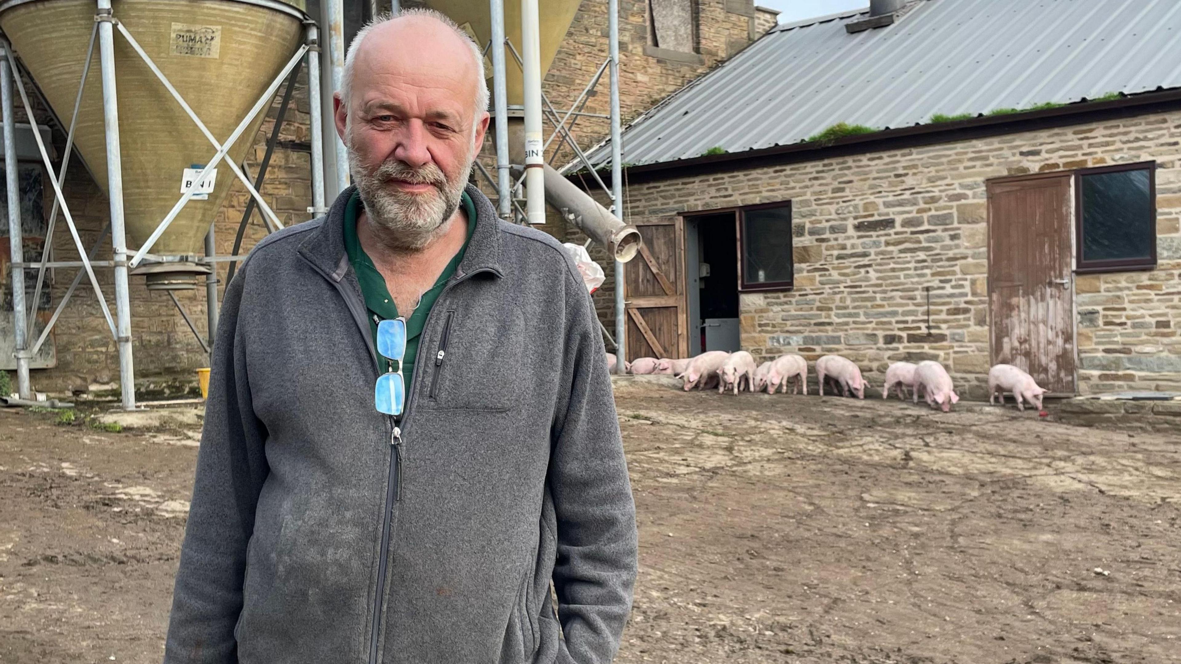 A balding man with a white beard is wearing a grey fleece with a pair of glasses hung from the zip. He is stood on a farm with a number of pigs in the background and farm buildings