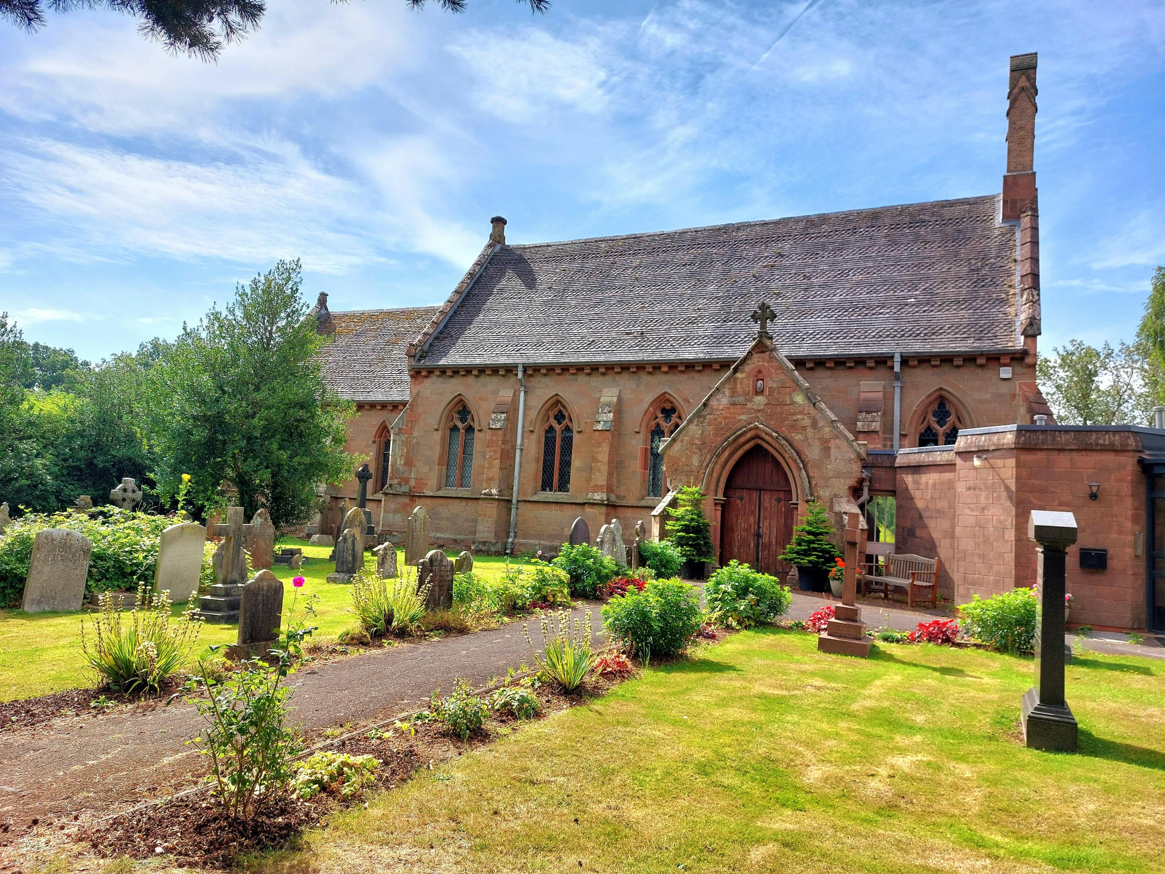 A church in salmon-coloured stone with arched windows and gravestones and flowers in the foreground