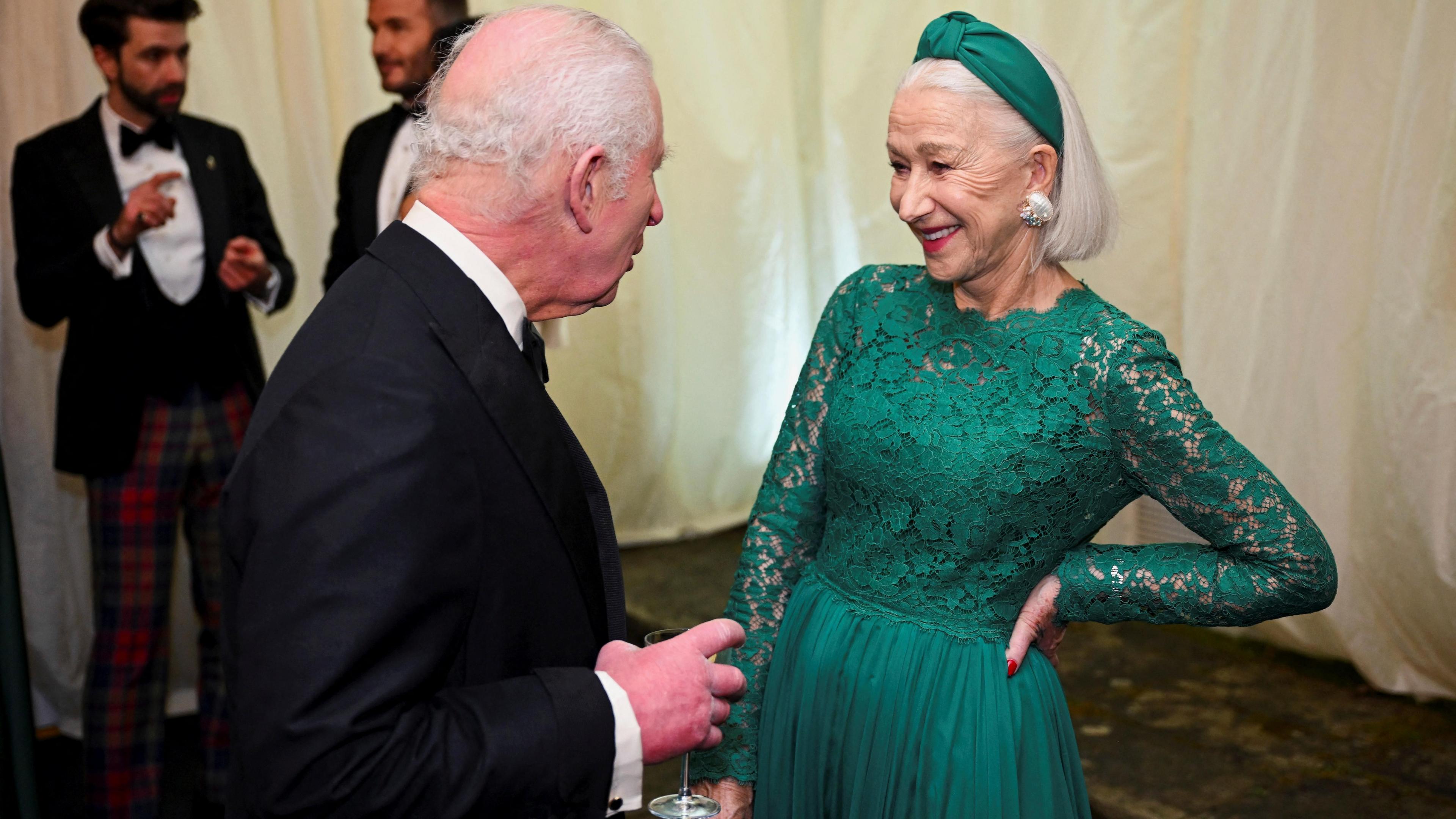 King Charles, wearing a black tuxedo, speaks with Dame Helen Mirren, who is wearing a long emerald green dress with a lace top, a matching green headband, and large silver earrings.