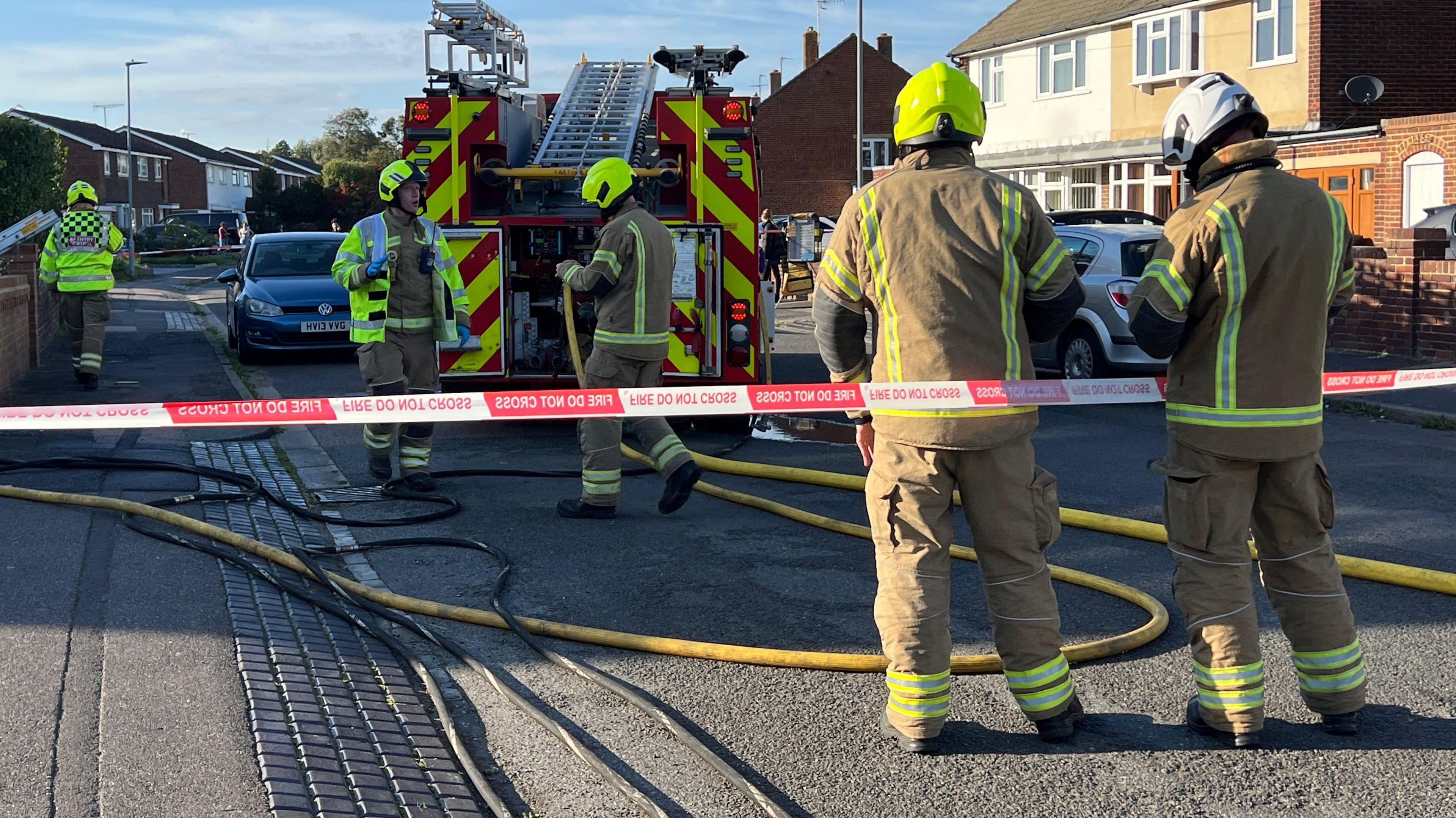 Firefighters standing on a residential road. There is a red and white cordon and a fire engine.