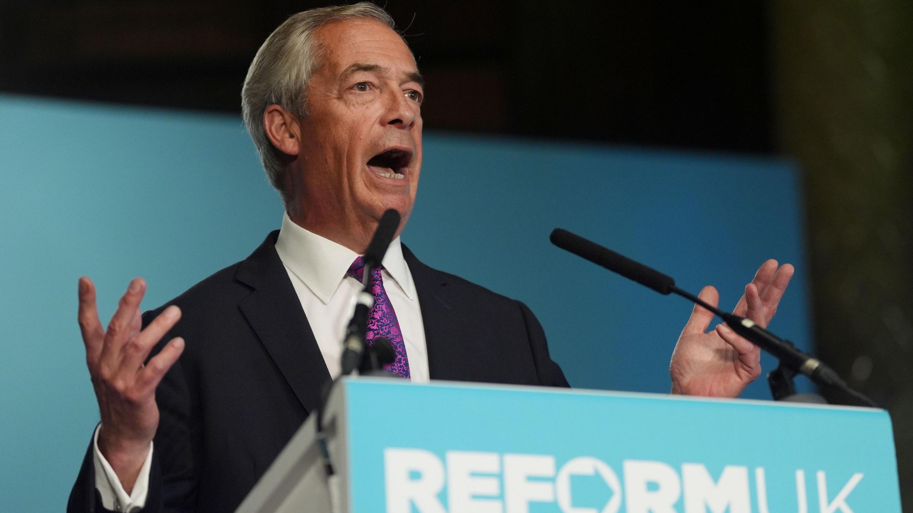Nigel Farage in a black suit and purple tie stands in front of a podium with "Reform UK" written on it, with its turquoise-coloured branding.