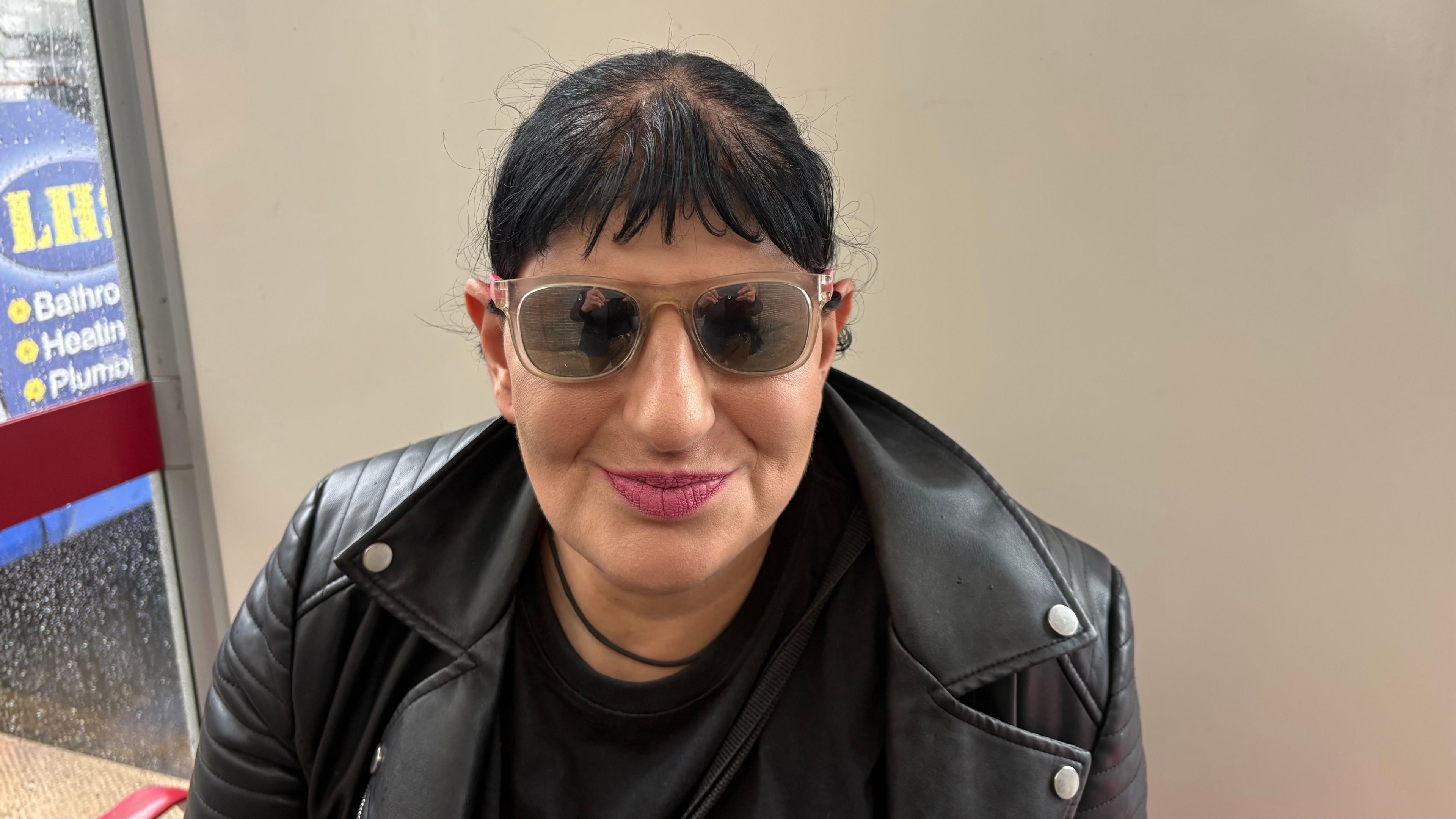 Troy Phillips with short black hair, wearing dark glasses and red lipstick. She has a black leather jacket over a black top and is sitting in a white-walled bus shelter. The back of a bus is visible to the left.