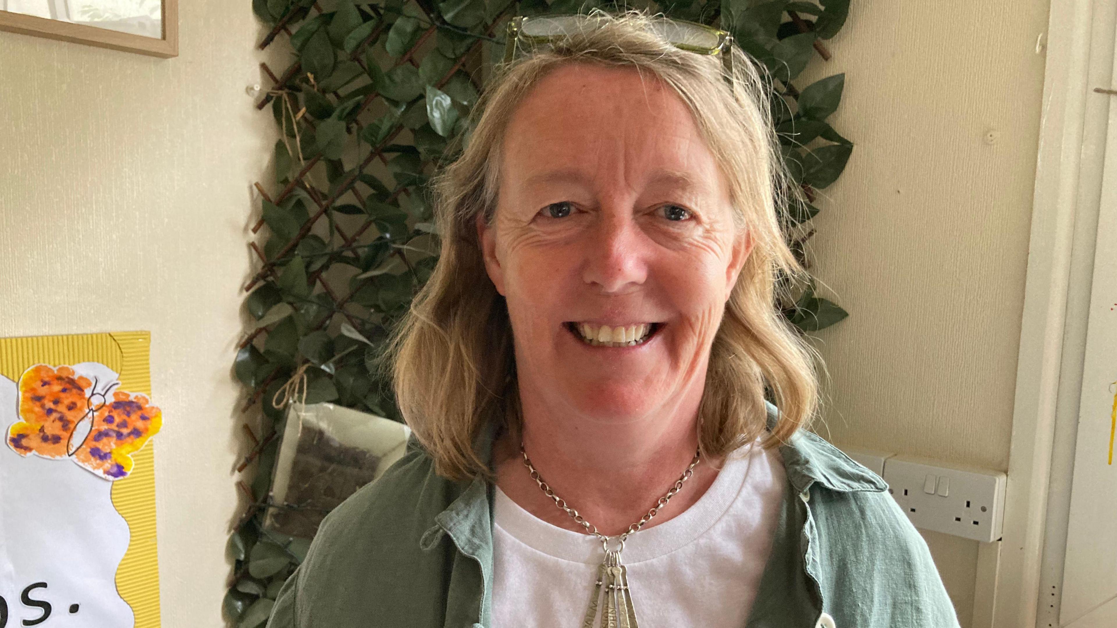 Beth Osborne pictured smiling at the camera with glasses pushed on top of her head. She wears a white t-shirt, a chunky silver necklace and a sage green overshirt. 