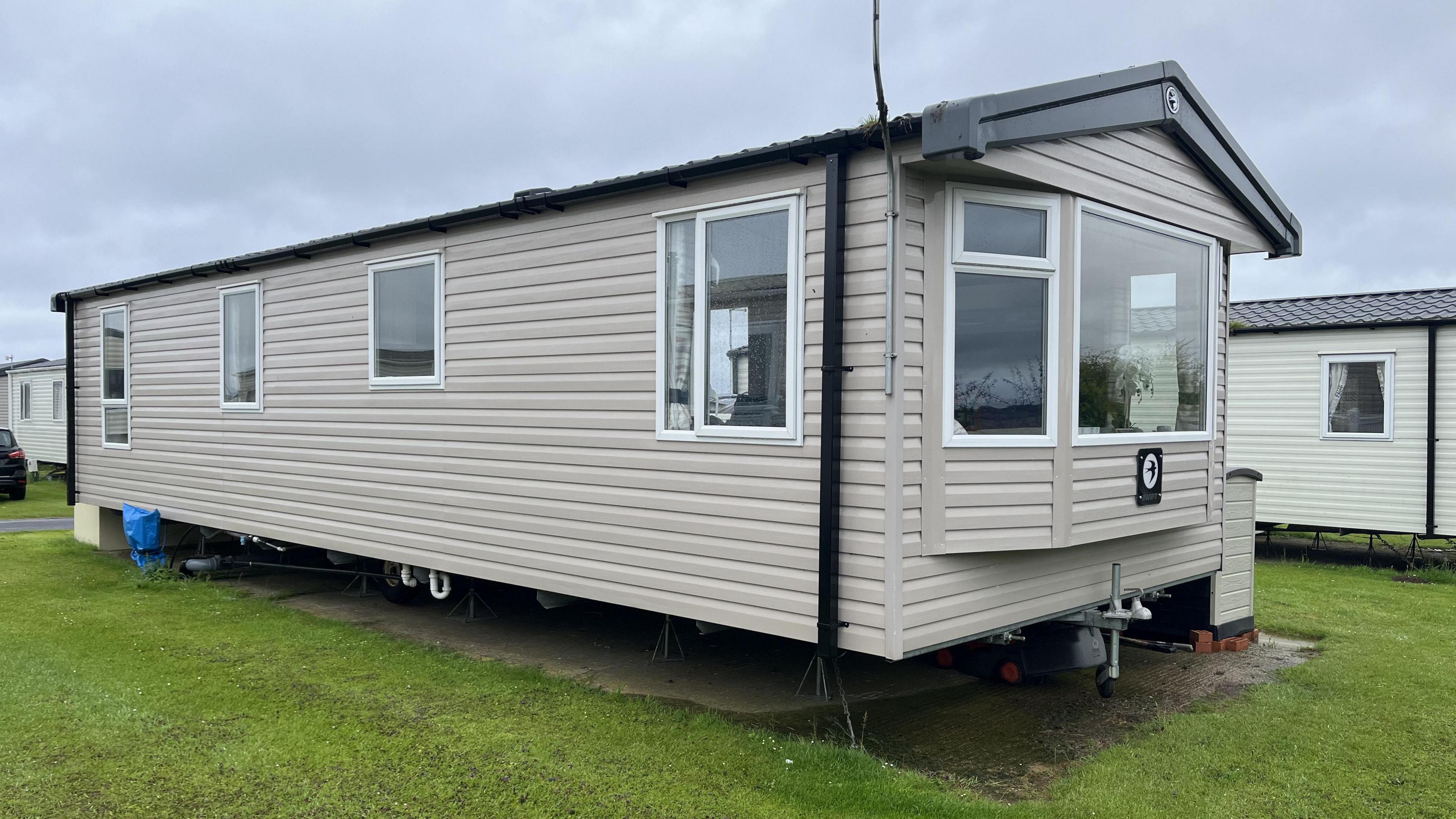 The Dawsons' caravan at Skipsea Sands holiday park. It is beige-coloured with white windows and black gutters and a bay window at the front. It is surrounded by grass and other caravans can be seen in the background.