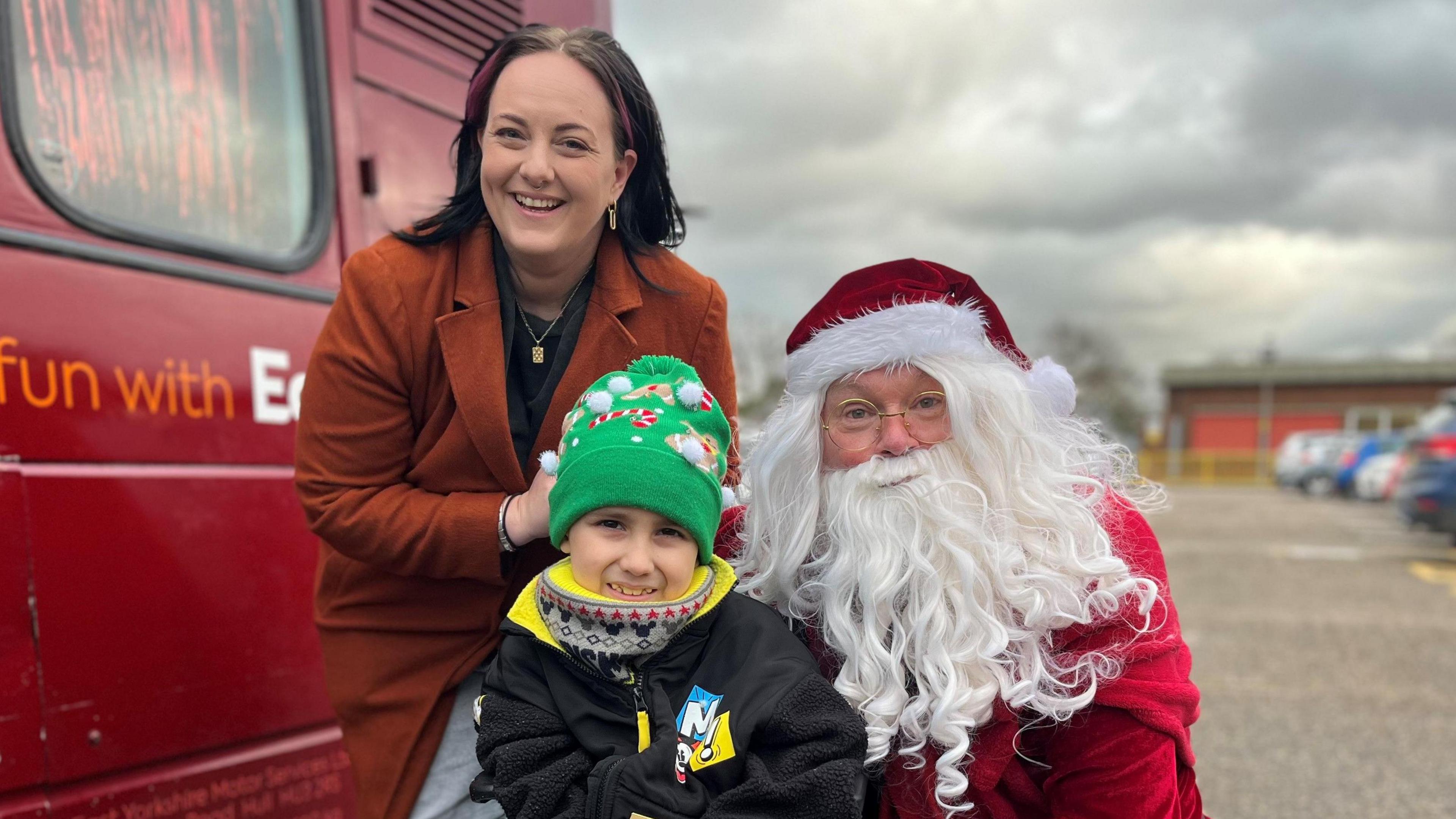 Isaac, his mum Jess and Santa posing by the Santa Bus. Isaac is wearing a black and yellow jacket with a green Christmas-themed bobble hat. Jess has dark hair with red higlights and is wearing a brown jacket. Santa is dressed just as you would expect for Santa. 