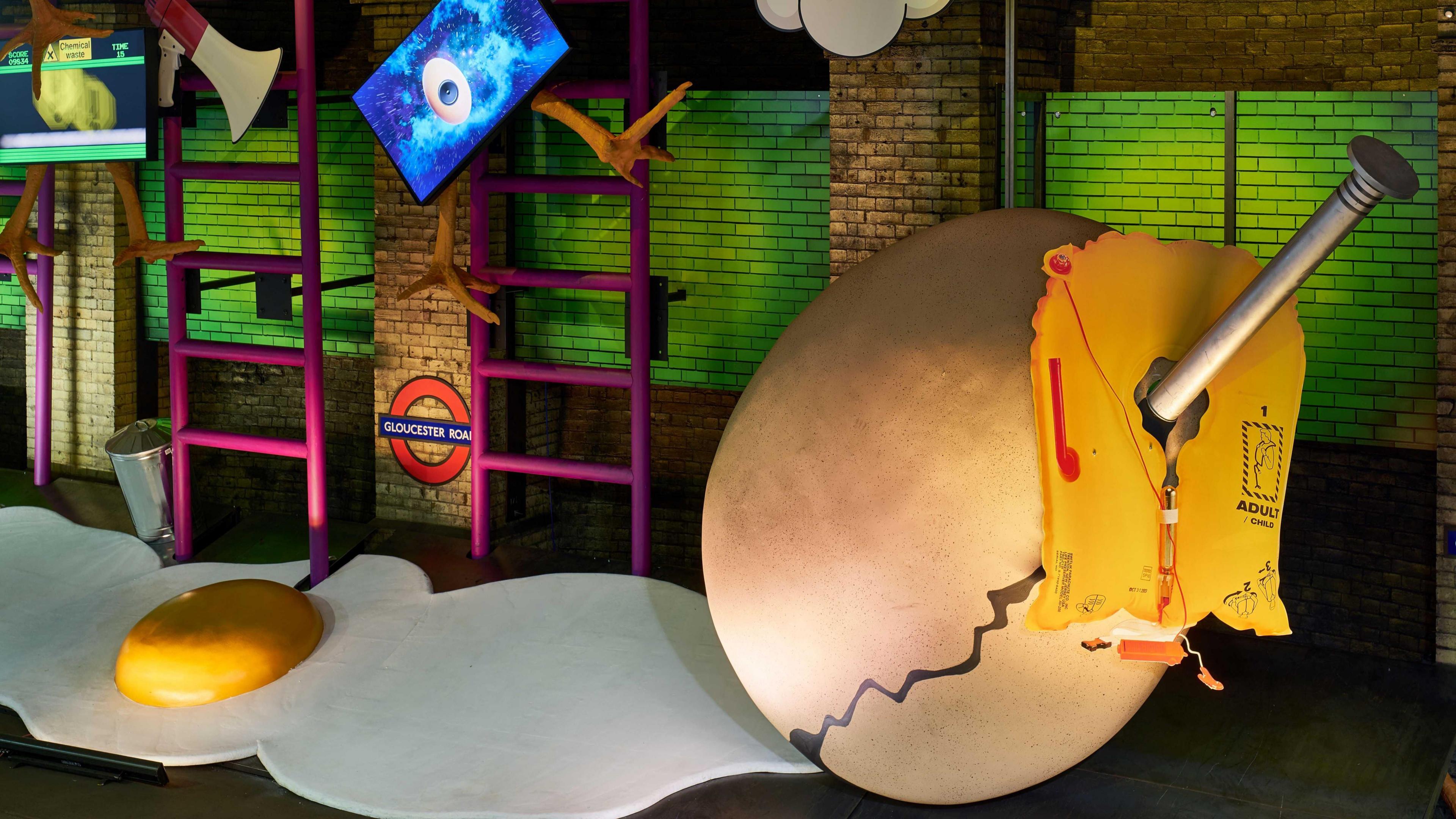 A surrealist art exhibit on a disused Tube platform featuring two giant cardboard eggs, one cracked open with a yellow yolk and the other with a nail sticking out of it. There are oversized ladders, megaphones and large chicken's legs as part of the exhibit. 