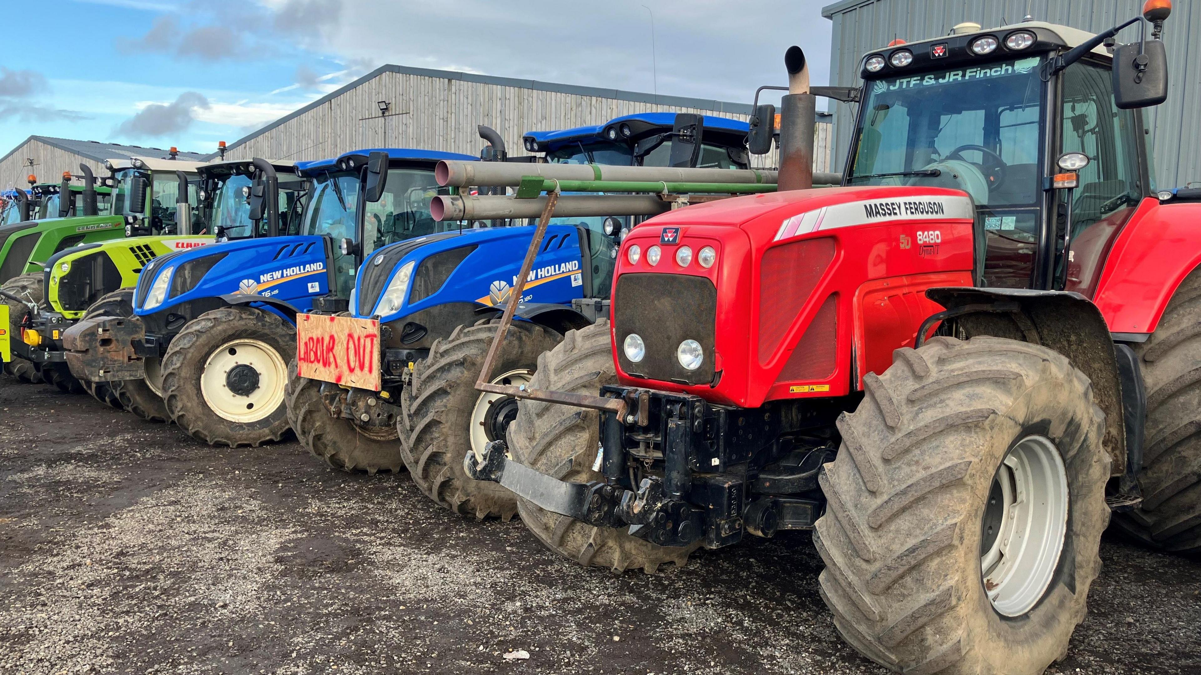 A row of tractors are lined up in parking spots. They are various colours including red, blue and green.