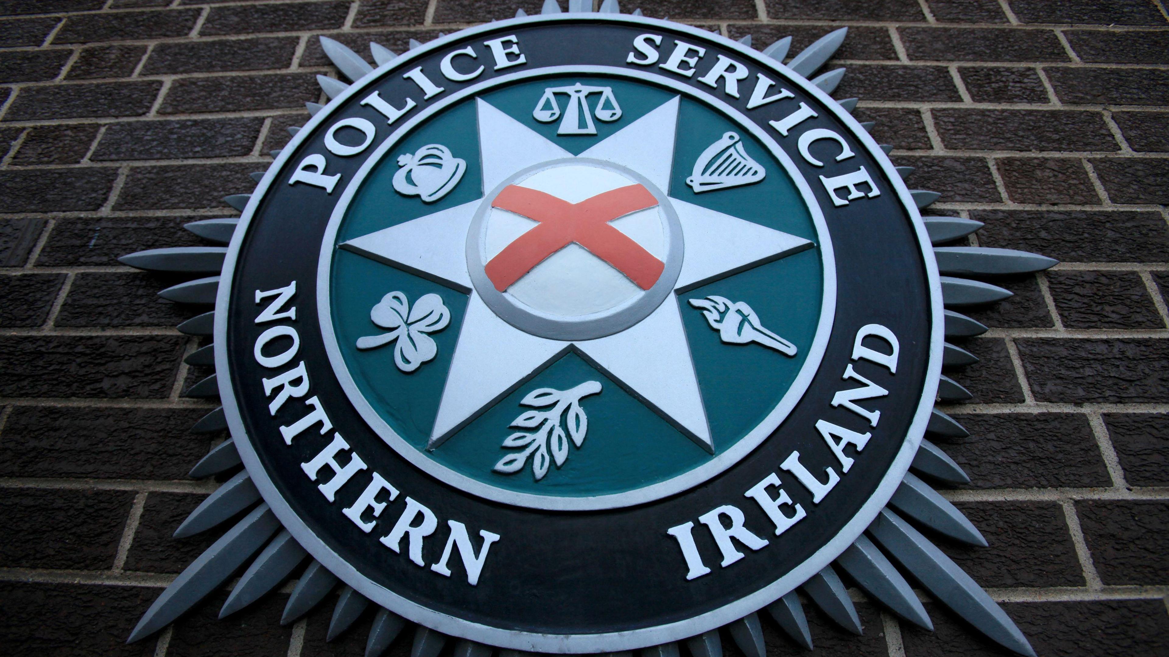 The PSNI emblem on a brick wall. It includes scales, a harp, a torch, a leaf, a shamrock an a crown.