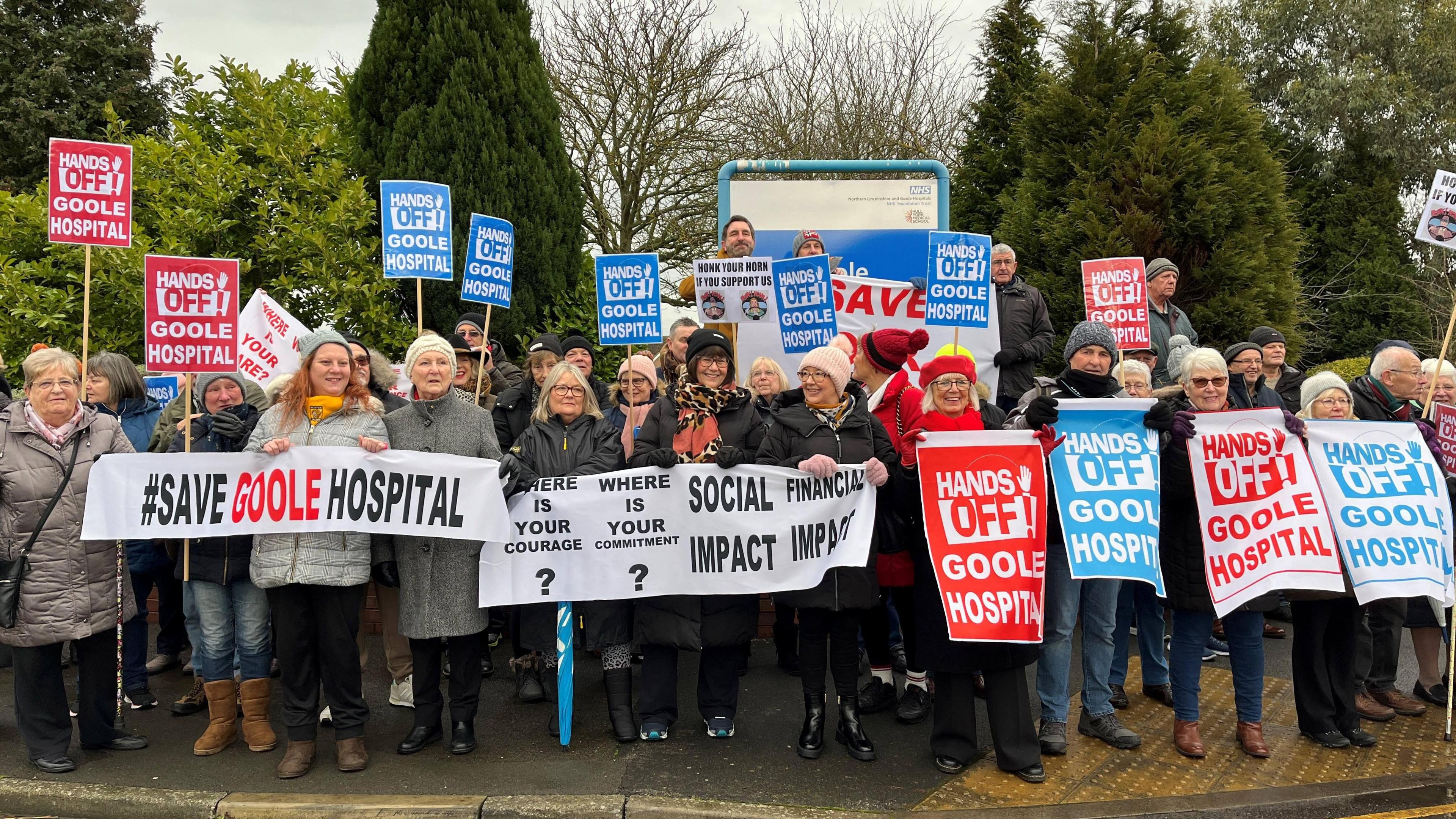 Group of people standing in front of trees holding placards, banners and signs with slogans about saving Goole hospital.