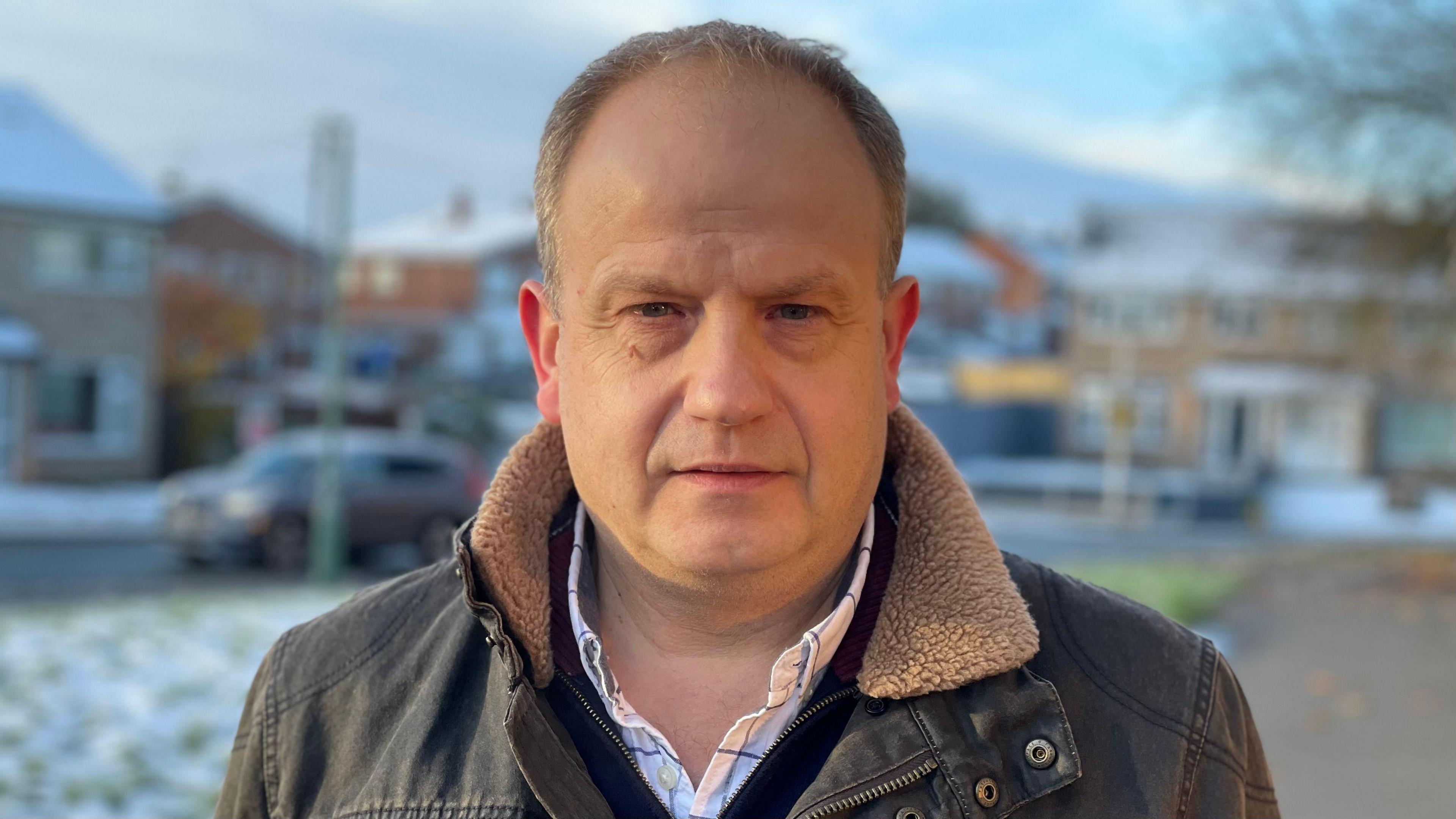 Councillor Dan Morris on a residential road, wearing a brown coat with snow on the ground behind him. He has short, light-brown hair and is clean shaven. A blurred image of a car next to a green verge is behind him with buildings, blurred, in the distance beyond.