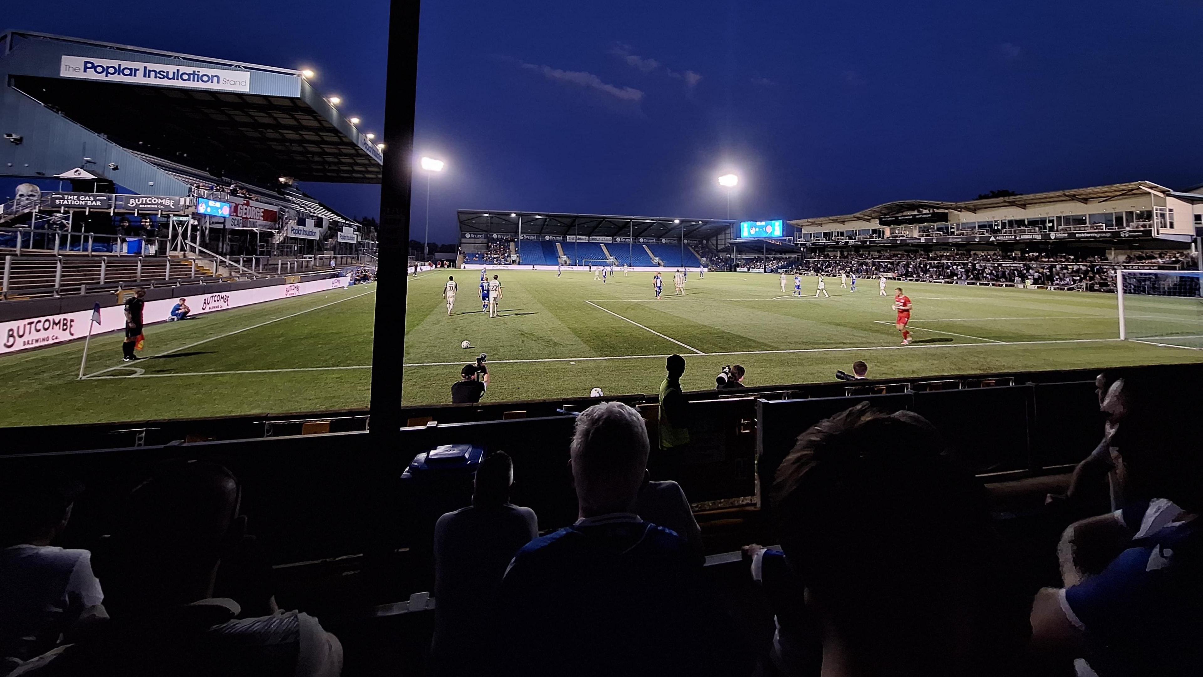 A view of the Memorial Stadium taken from the terracing at one end during an evening match between Bristol Rovers and Cambridge United. The floodlights are on and spectators are visible in three of the stands but not in the stand at the other end of the pitch