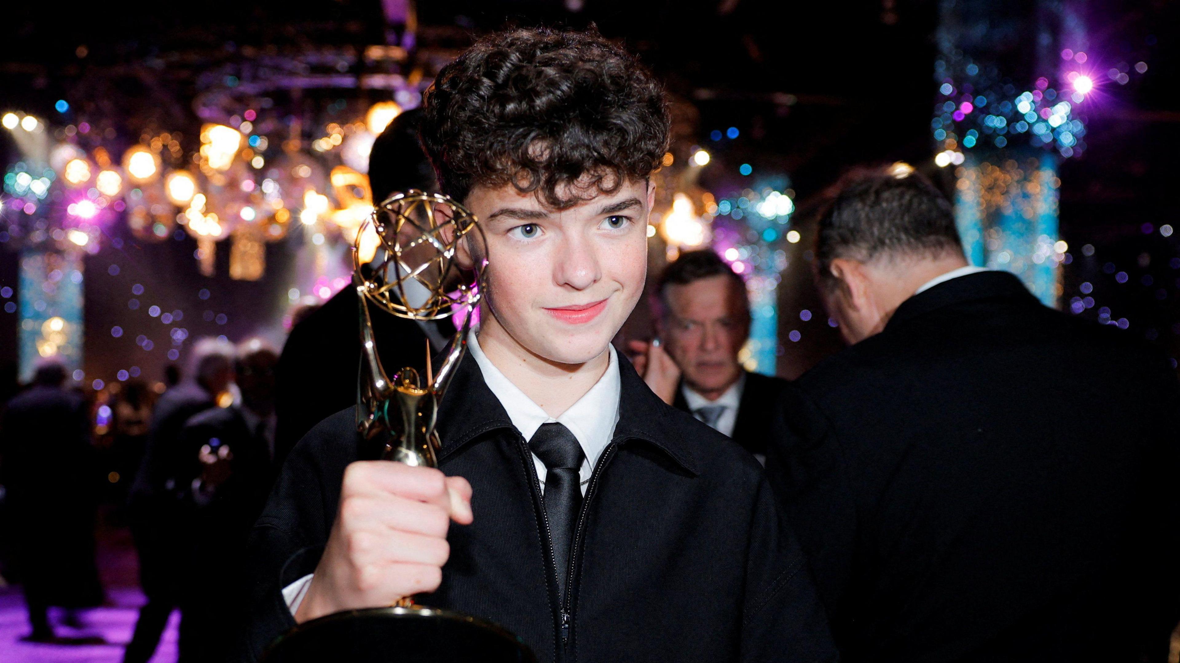 Owen Cooper holds up his Emmy award on the purple carpet. He wears a suit with a black tie and white shirt. There are yellow, blue and purple lights hanging from the ceiling behind him.