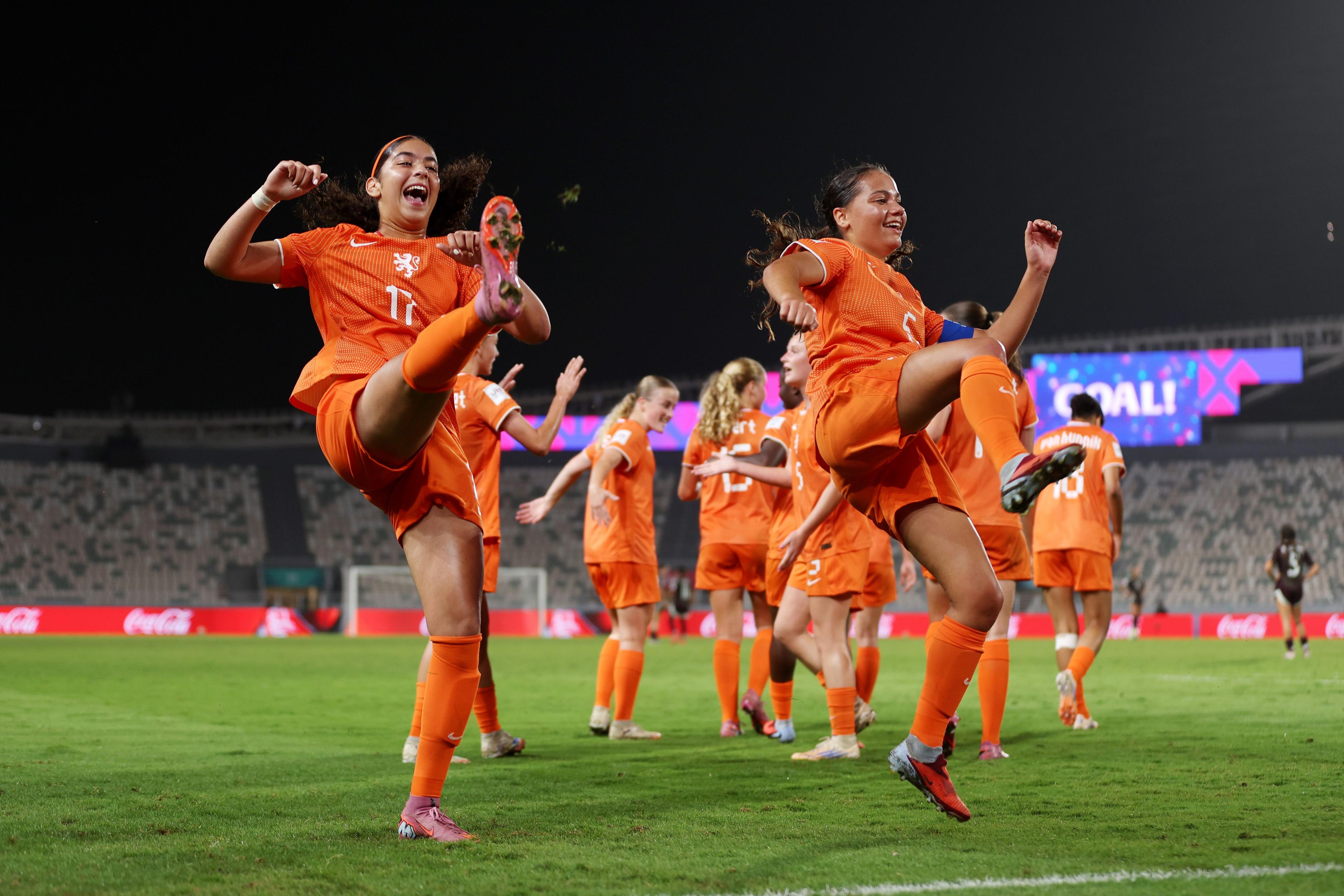 Football players in bright orange uniforms performing high kicks during a warm-up on a green field, with teammates in the background and a stadium lit by bright lights and colorful advertising boards under a dark night sky.