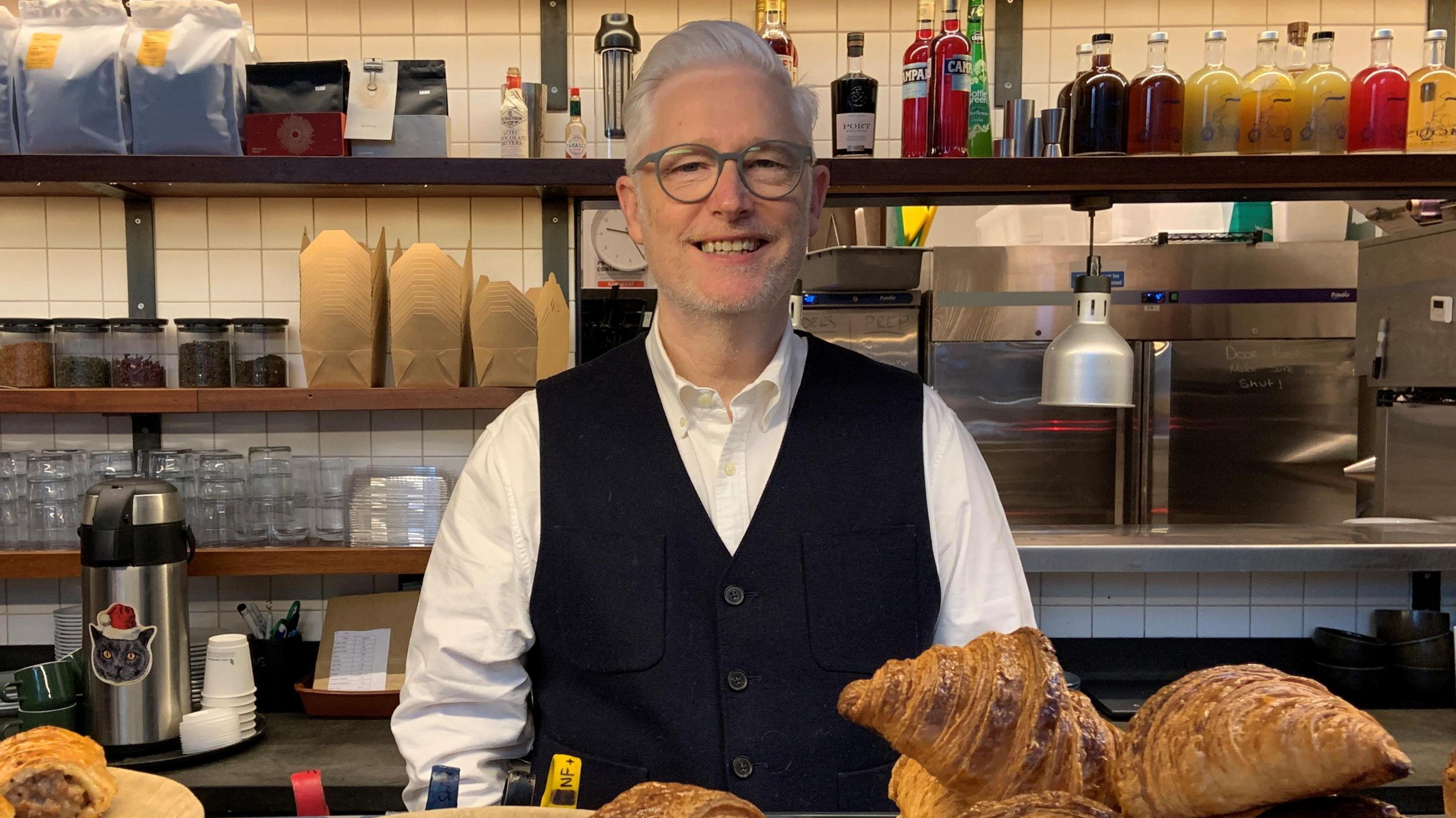 Photo shows a smiling man with white hair and glasses in a white shirt and black waistcoat. He is stood behind the counter in a coffee shop. There is coffee equipment and drinks on shelves behind him and a counter full of pastries in front of him
