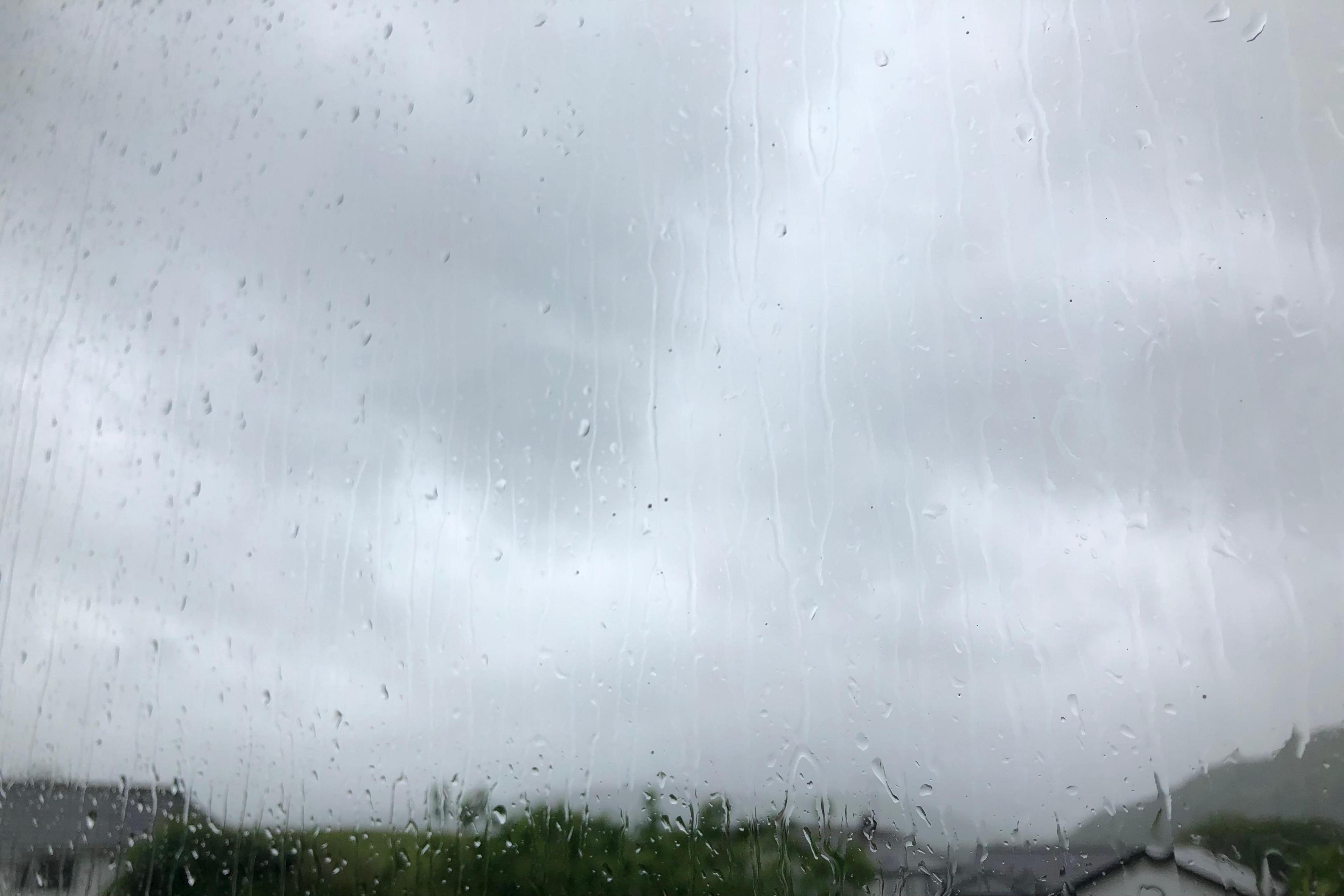 Photo taken through a window which has lots of rain streaks on it. There are massive grey clouds in the sky and the tops of houses and trees are just visible.