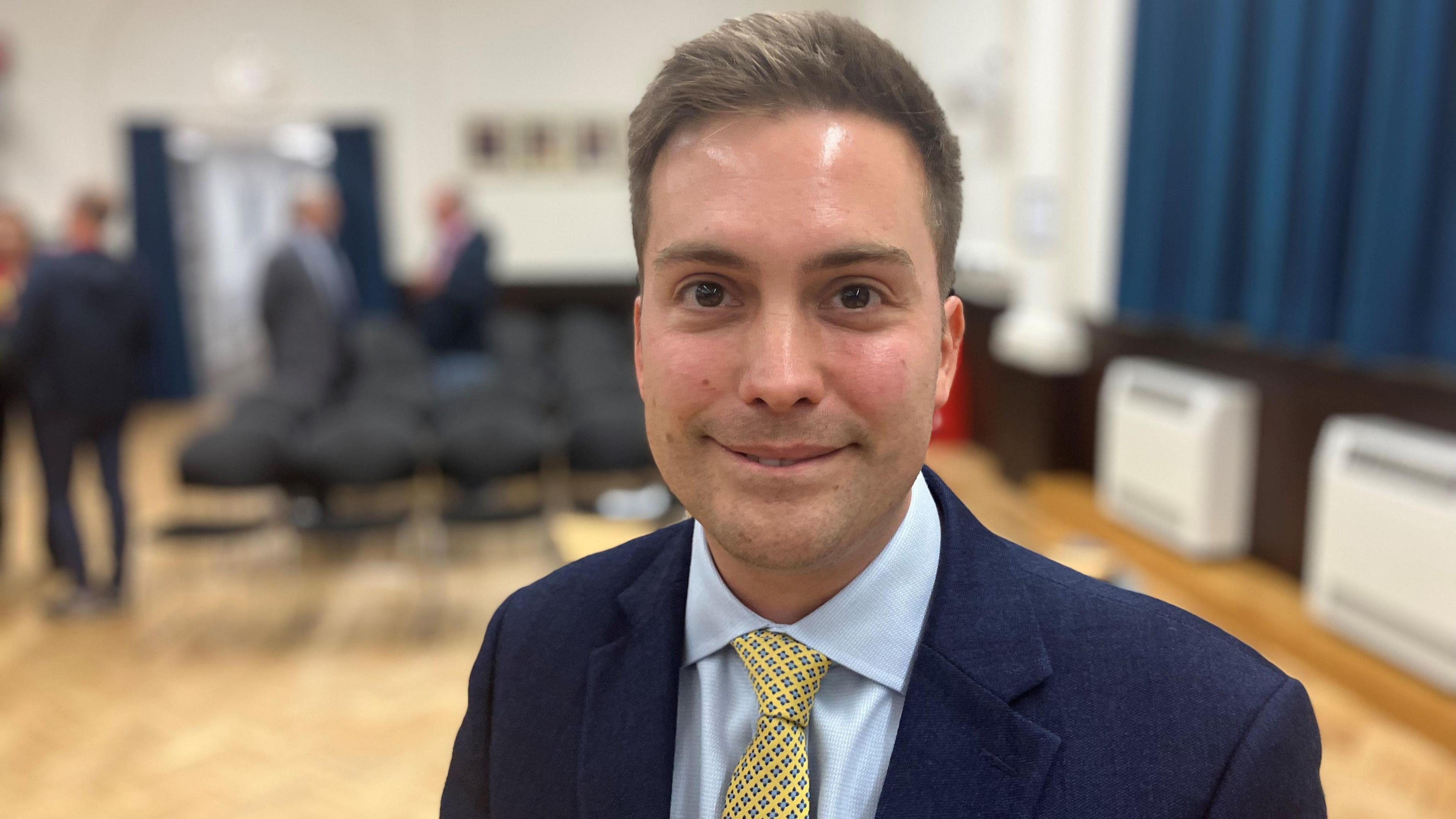 Rob Hunter, dressed in a blue suit, light coloured shirt and yellow tie, stands in a hall, looking at the camera