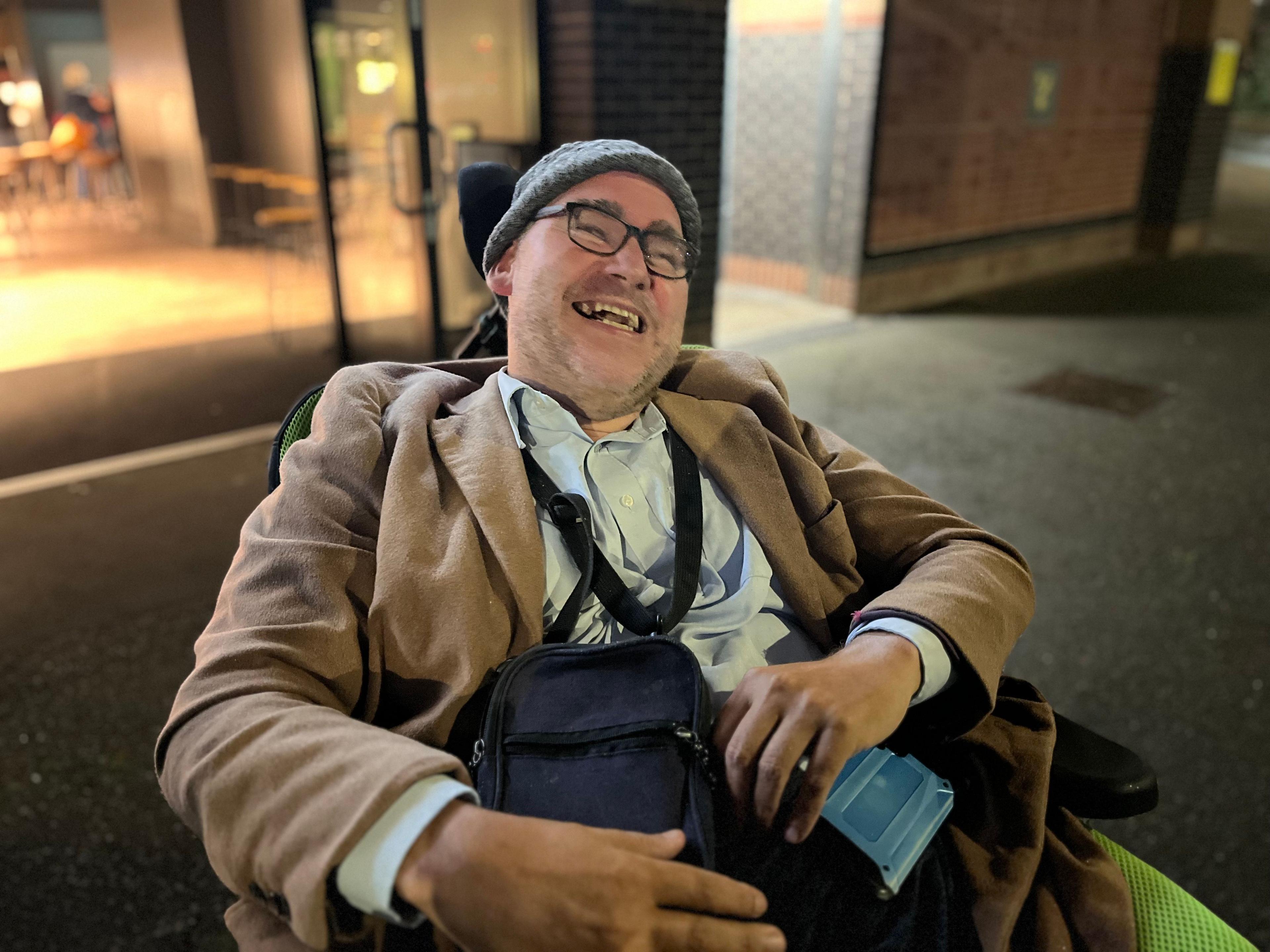 A man leaning back in a wheelchair and smiling, outside at night. He is wearing a grey woolly hat, a brown jacket, pale blue shirt and has a black bag on his lap