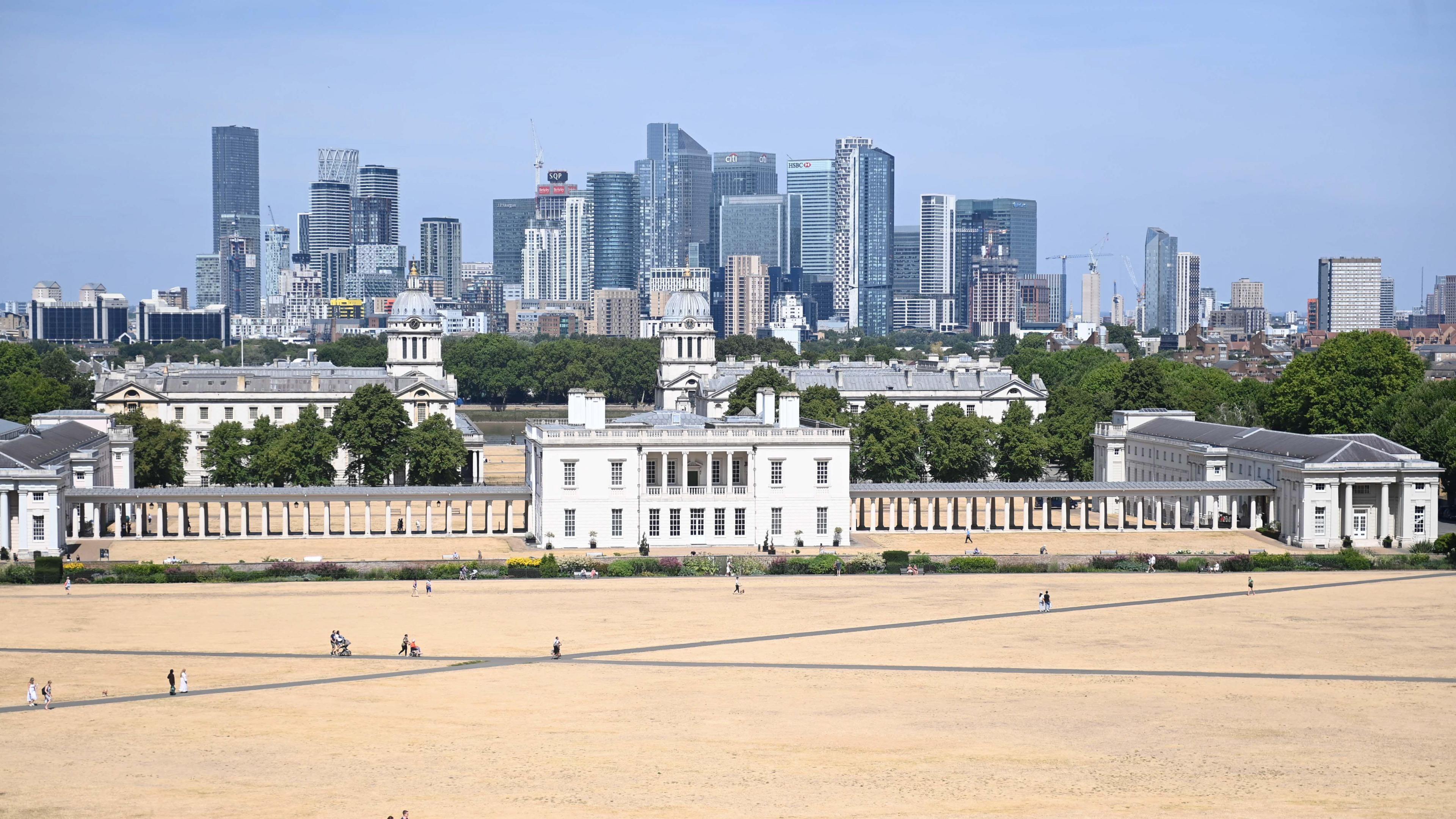 A hot-looking, dry, parched park with skyscrapers in the background with a clear blue sky overhead