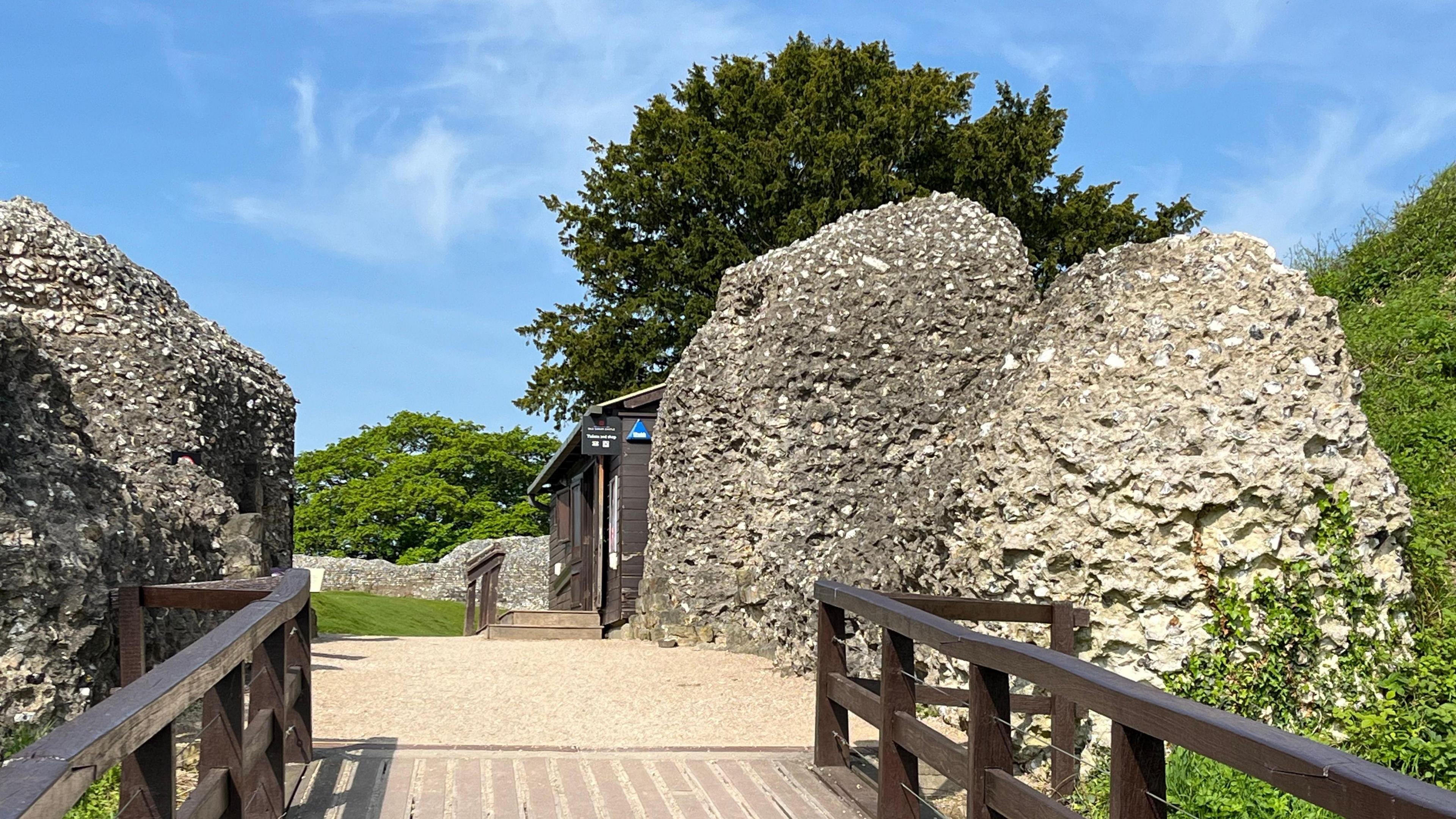 View from a footbridge going into ruins and a visitors wooden hut. Green grass, trees and blue skies.