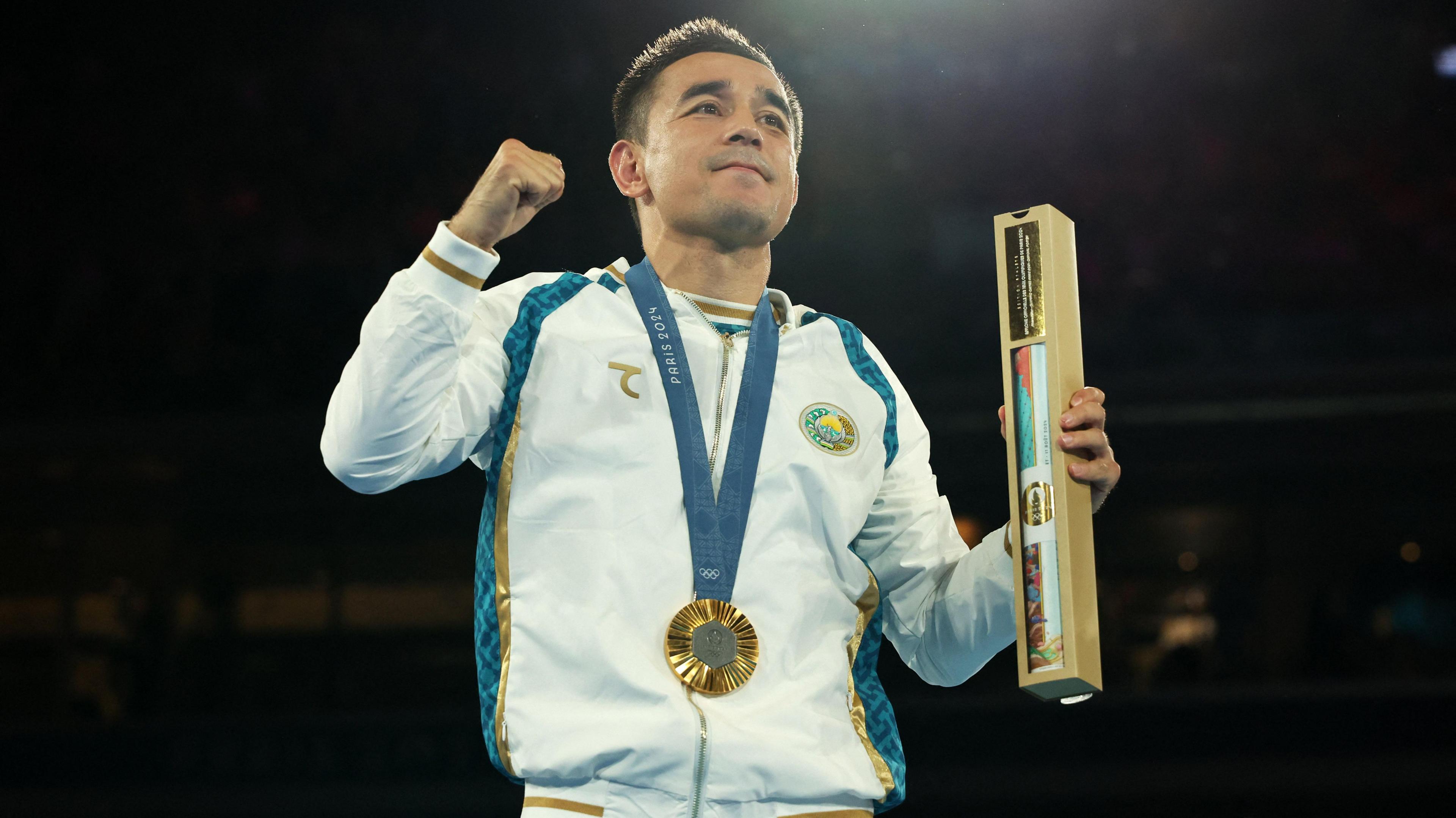 Hasanboy Dusmatov holds his fist in the air celebrating with an Paris 2024 Olympic medal hanging round his neck. He is wearing a white tracksuit with blue and gold stripes