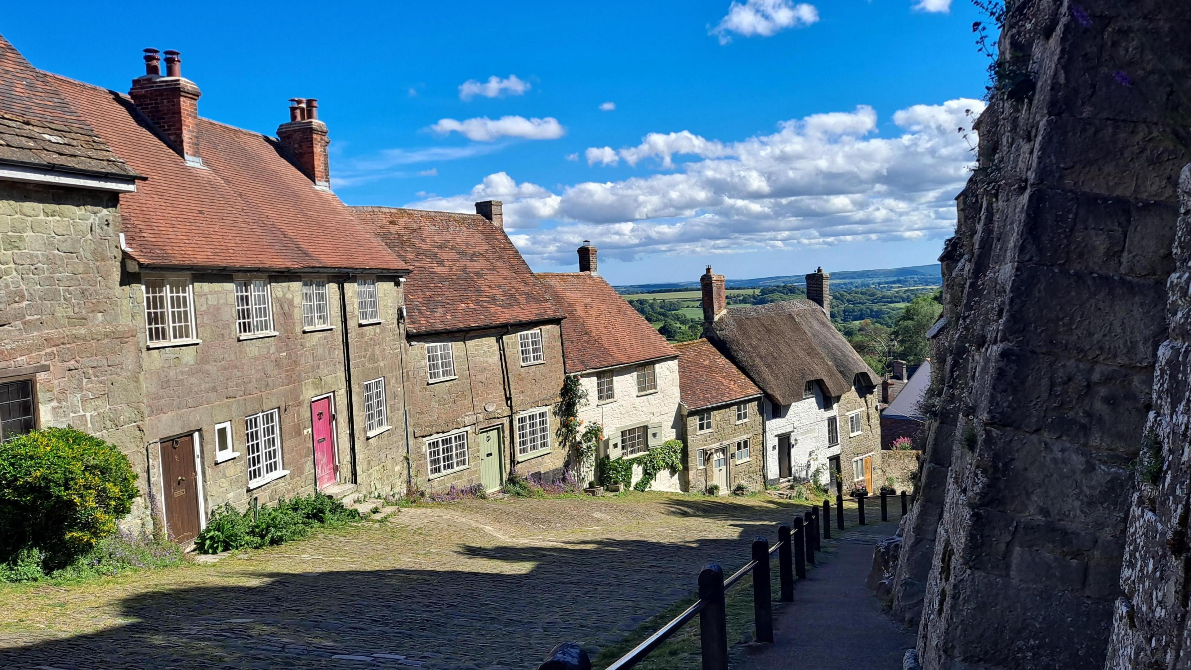 A steep cobbled street, with a row of historic houses descending on one side, and an old brick wall on the other. The countryside is in the distance.
