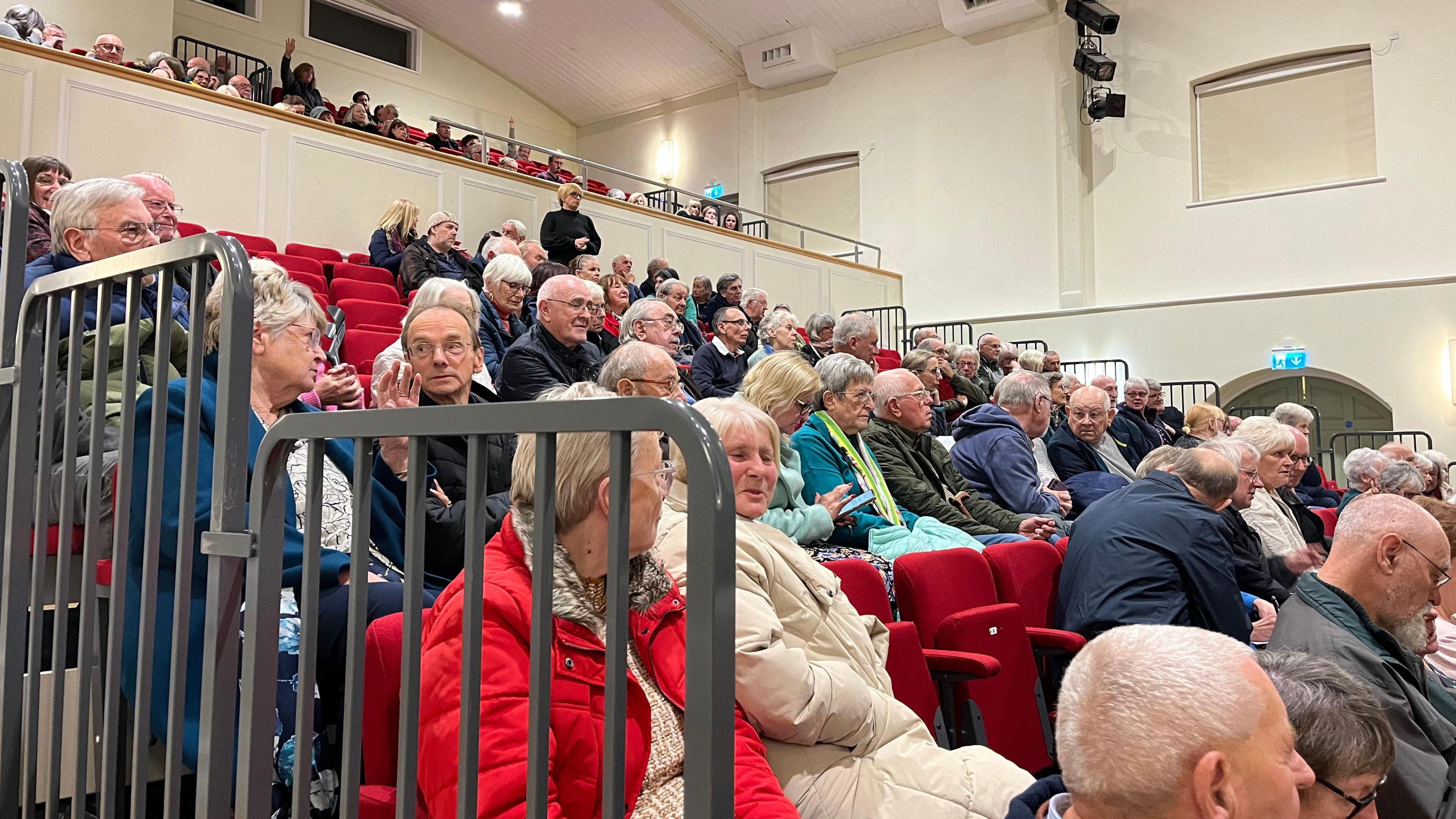 A crowd of people sitting in seats on a stand, they are speaking to each other or looking towards the front of the room. 
