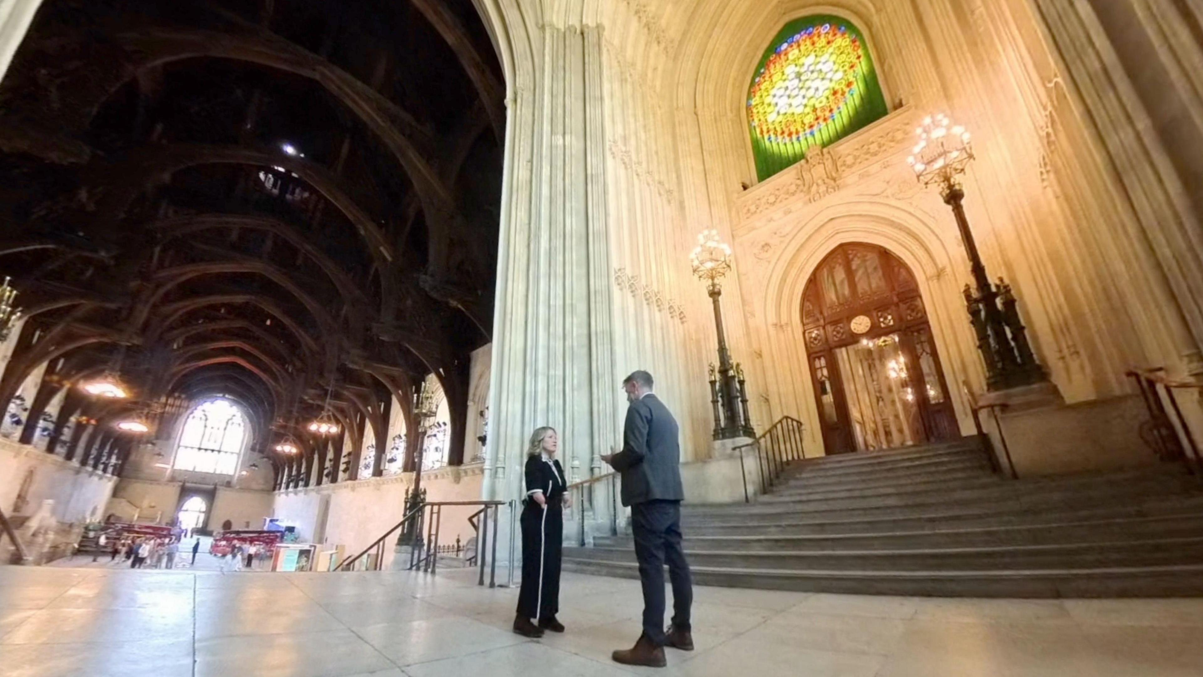 A wide-angled image of a man in a grey suit and a woman in a navy suit stood talking to each other on a mezzanine between two sets of stairs. A large hall on the lower floor, with a high wooden vaulted ceiling and large stained glass window at the far end. A set of stone steps leads up to the first floor on the right hand side. There are two ornate lamps at the top of the steps on either side of an ornate wooden door. Above the door is a stained glass window.