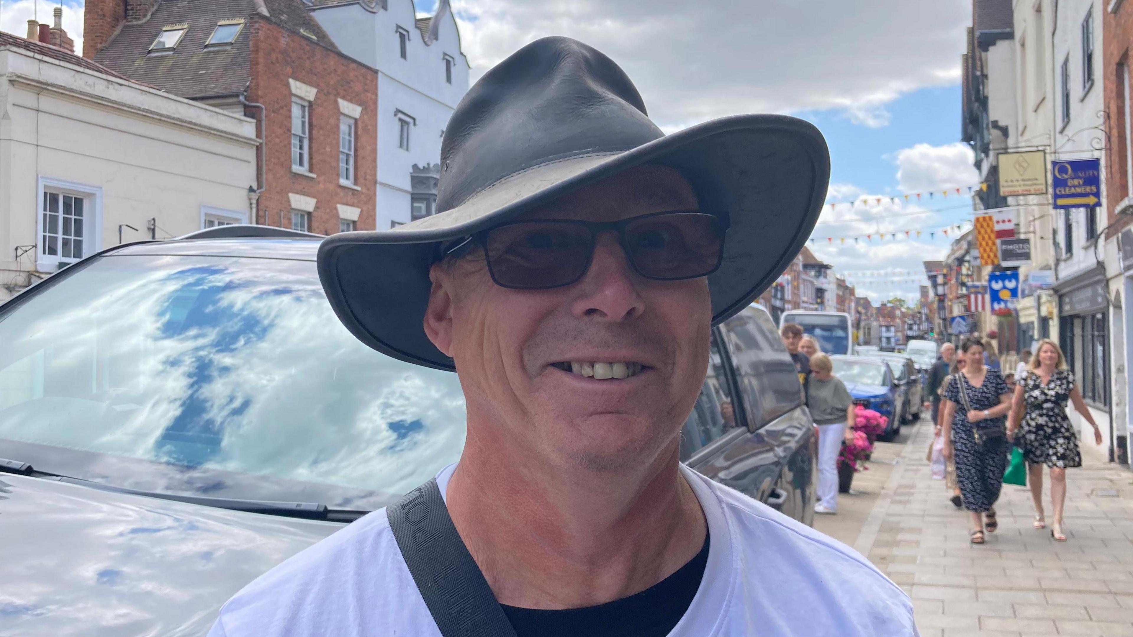 A photo of a white man wearing sun glasses and a wide brimmed hat and a white t-shirt. He is stood in front of a car with a street to his right, with two women walking towards him