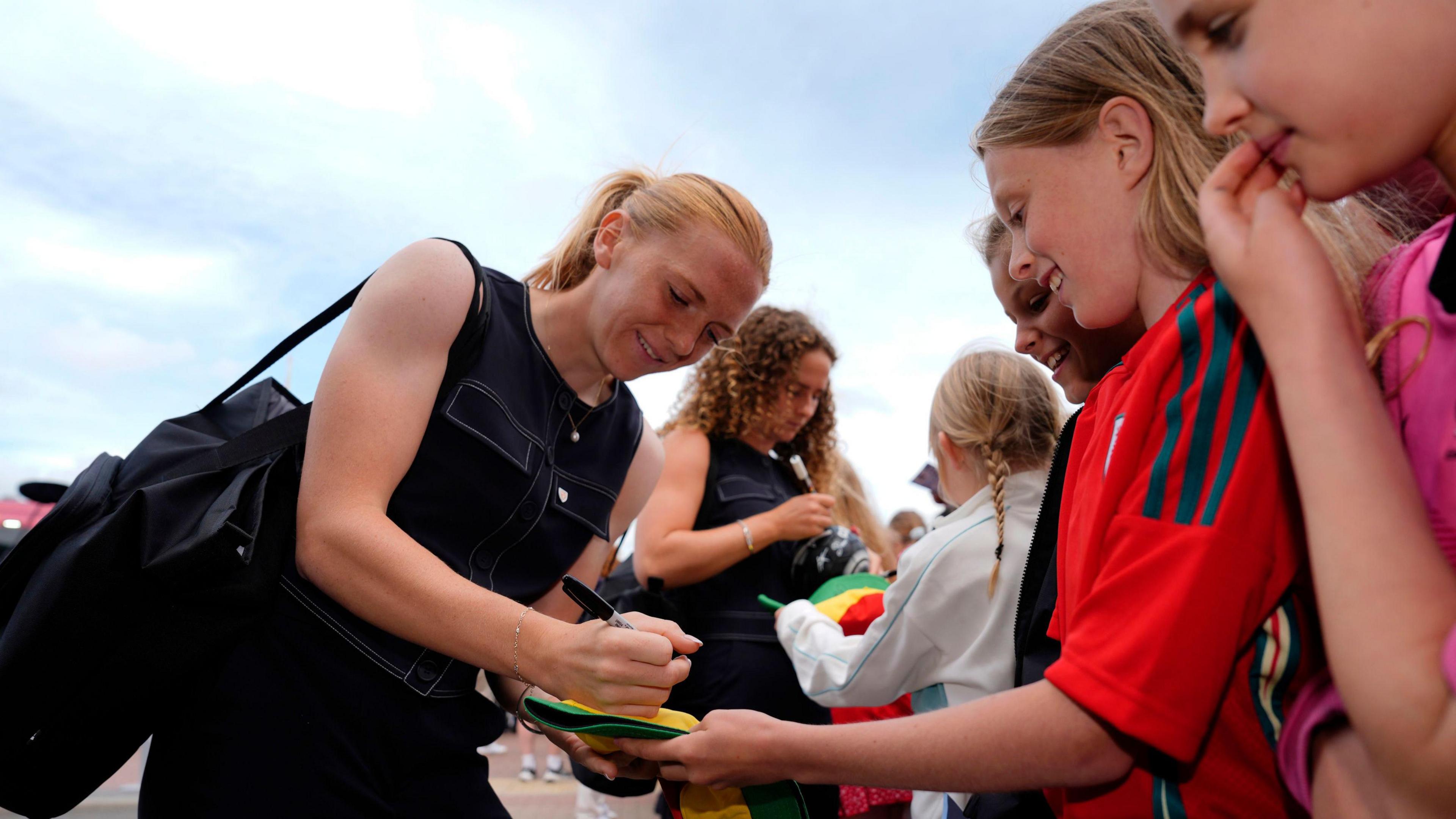 Wales' Ceri Holland wears a black sleeveless top and dark trousers. She is signing an autograph for young fans on the right