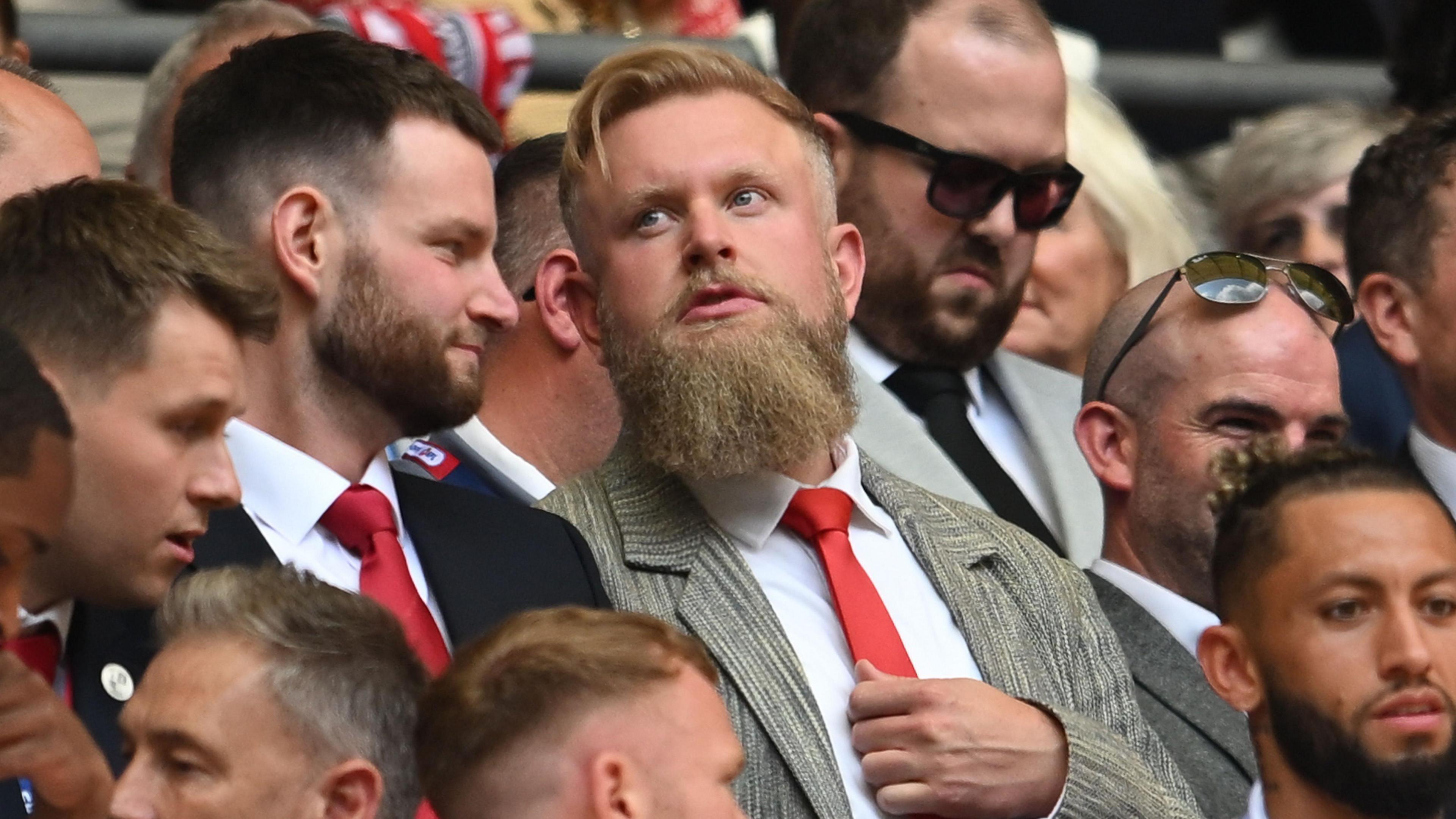 Preston Johnson in a grey suit, white shirt and red tie, sitting in the crowd at a Crawley Town football match