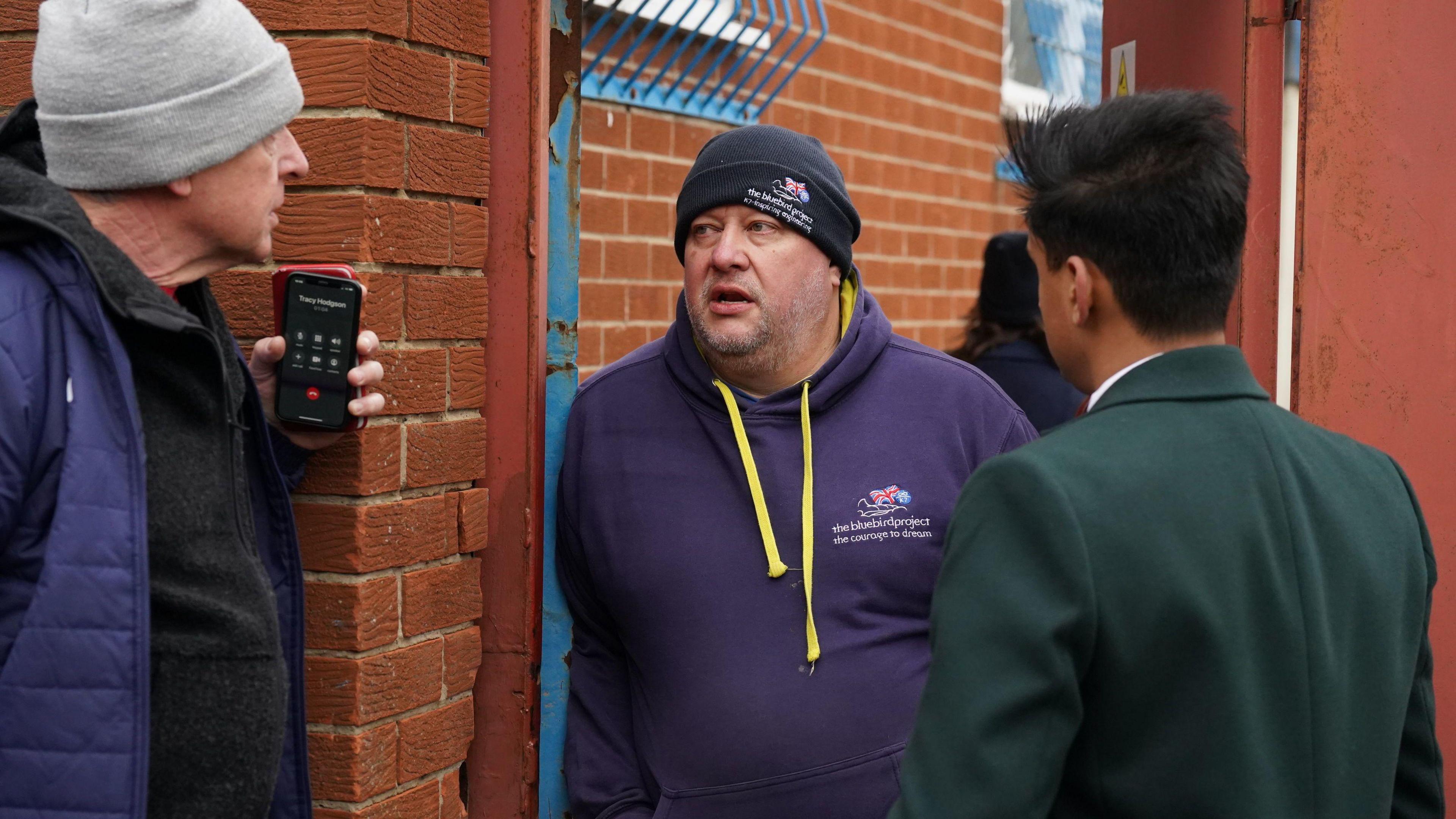 Bill Smith (centre) talks to two men at the gate of his engineering yard in North Shields. He is wearing a blue woolie hat bearing The Bluebird Project logo as well as a blue hoodie with the same logo.