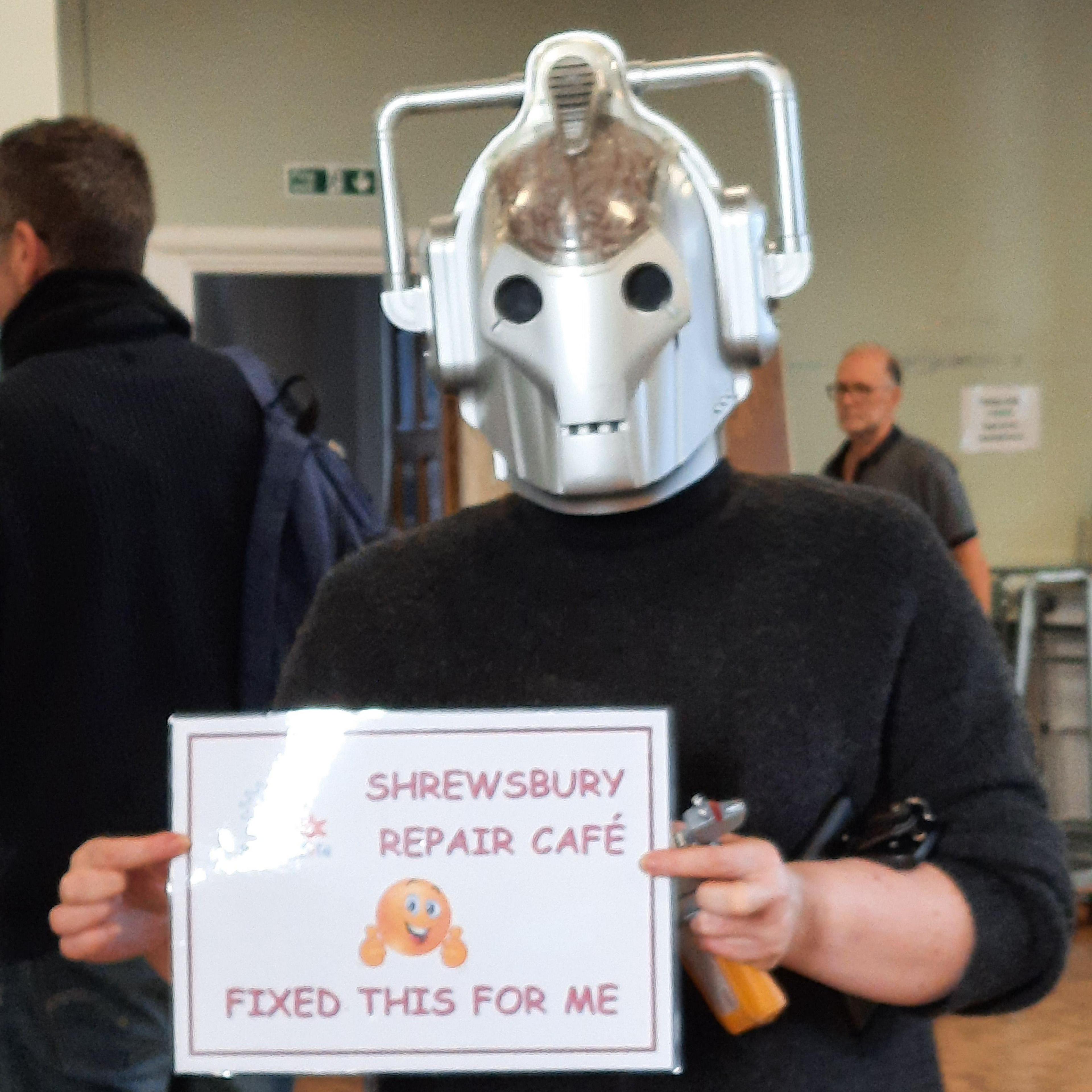 A person wearing a Cyberman helmet made of silver metal with the image of a brain in it. They wear a black jumper and hold a white laminated piece of paper which says 'Shrewsbury Repair Cafe fixed this for me' with an emoji of a face with two thumbs up on it.